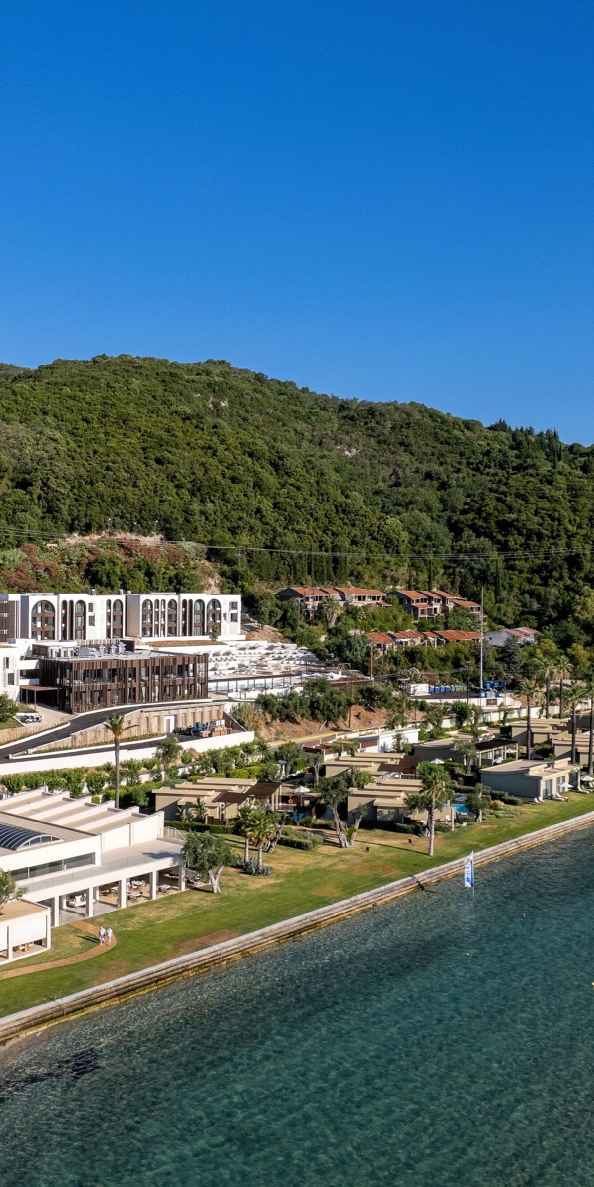 An aerial view of a resort's white buildings and low villas set among lawns and trees at the foot of a wooded hillside, with the sea in the foreground.