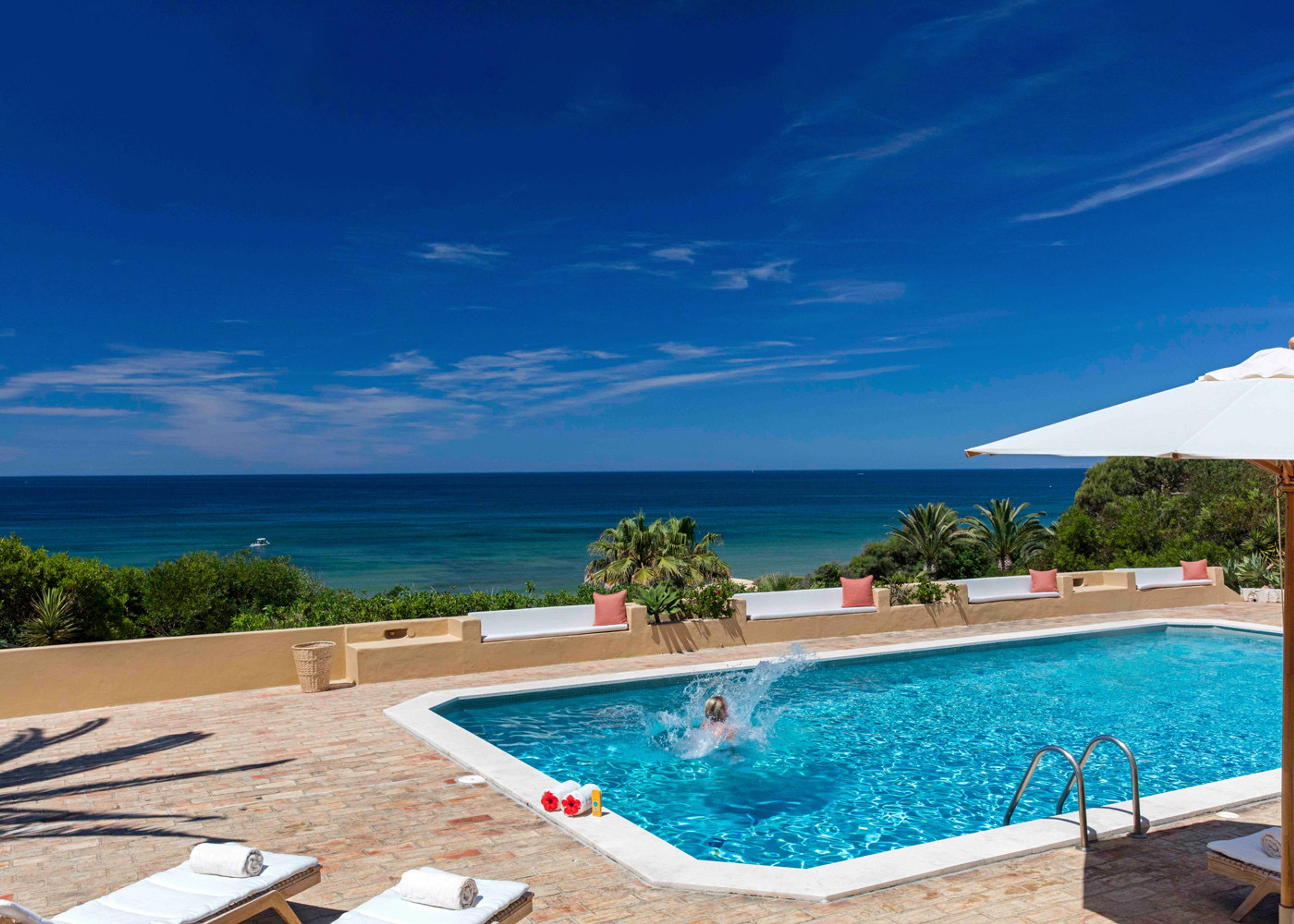 A person jumps into a swimming pool creating a splash, with sun loungers and the sea visible beyond the terrace.