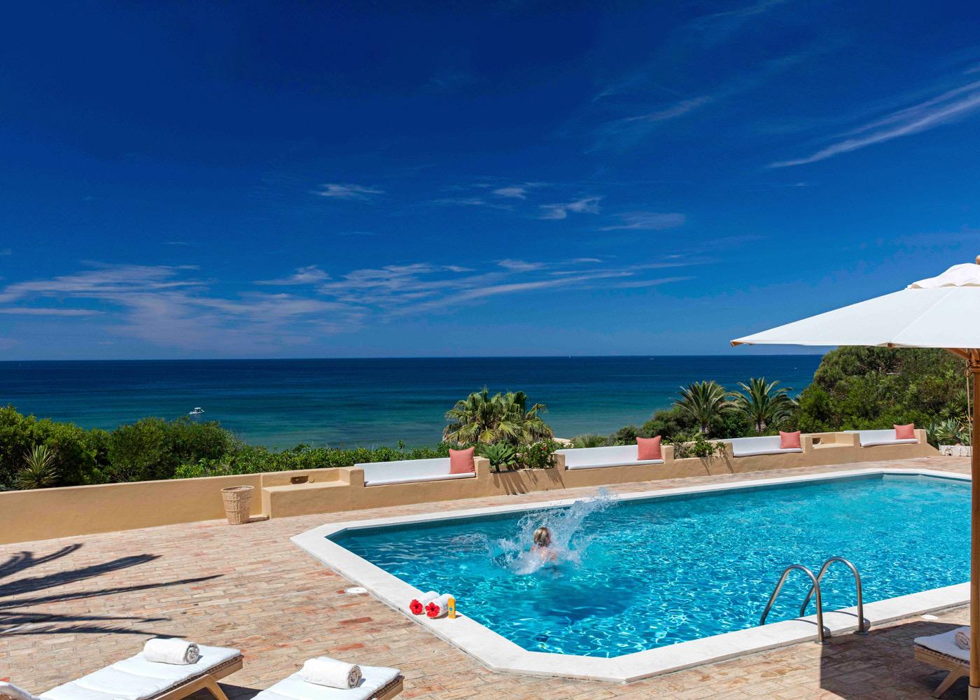 A person jumps into a swimming pool creating a splash, with sun loungers and the sea visible beyond the terrace.
