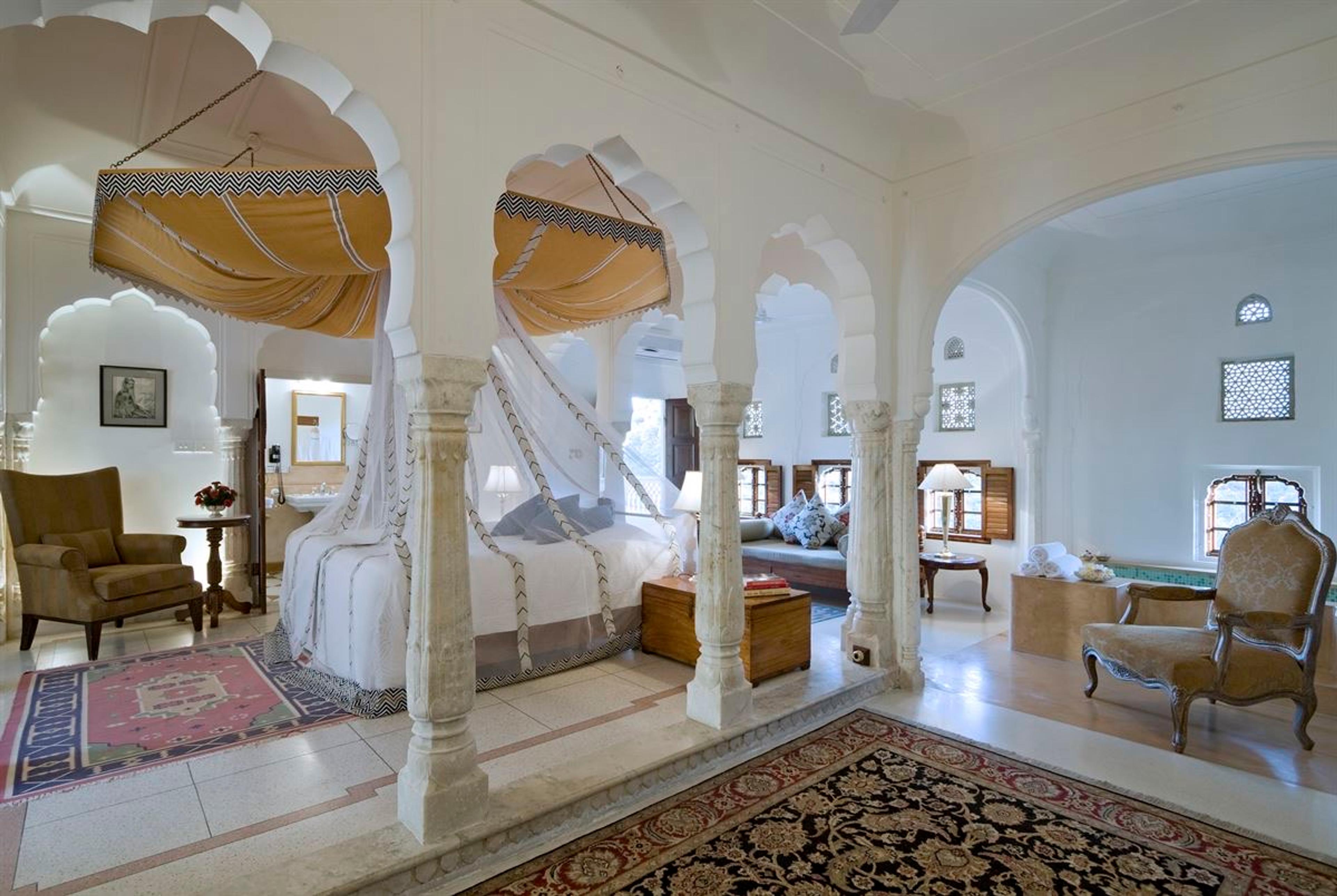 A suite with two draped canopy beds under carved stone arches, opening through columns to a sitting area and latticed windows.
