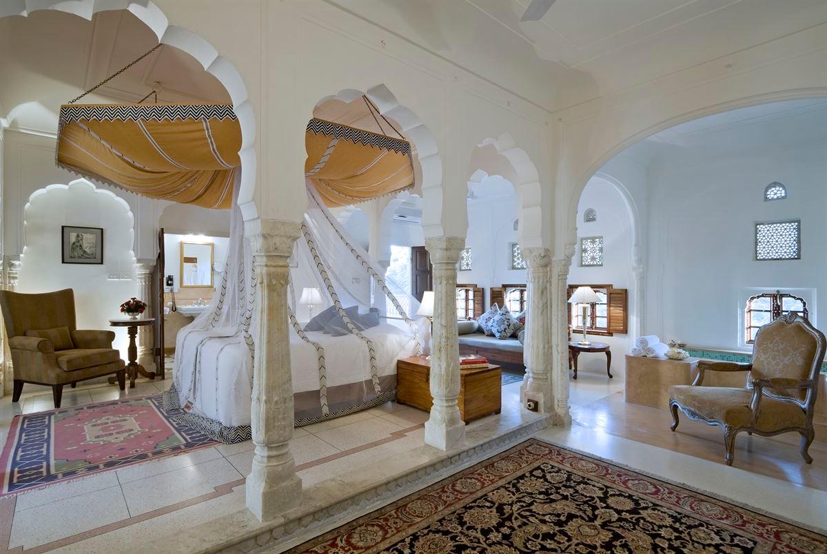 A suite with two draped canopy beds under carved stone arches, opening through columns to a sitting area and latticed windows.