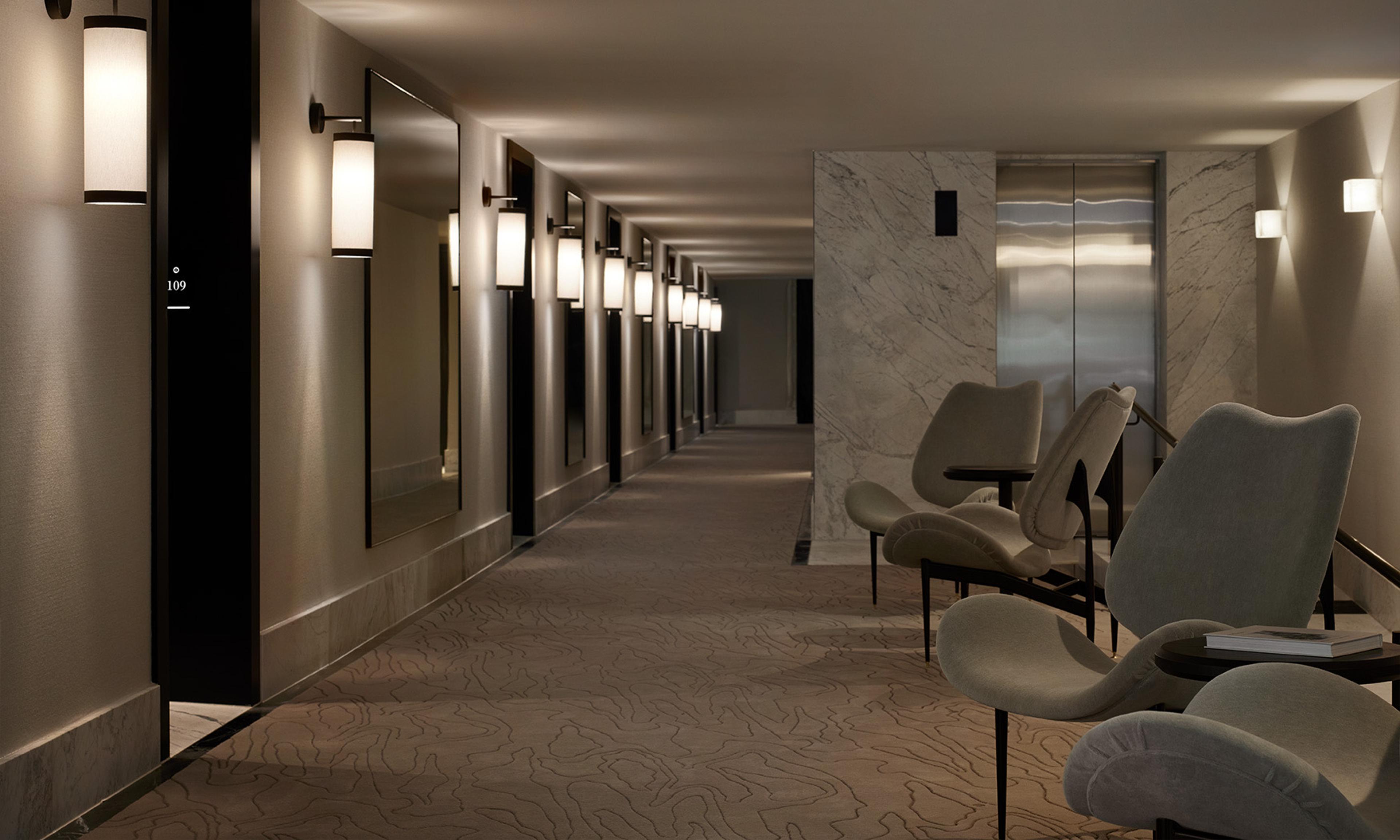 A long hotel corridor with wall sconces, framed mirrors, a marble lift lobby and upholstered chairs at the far end.