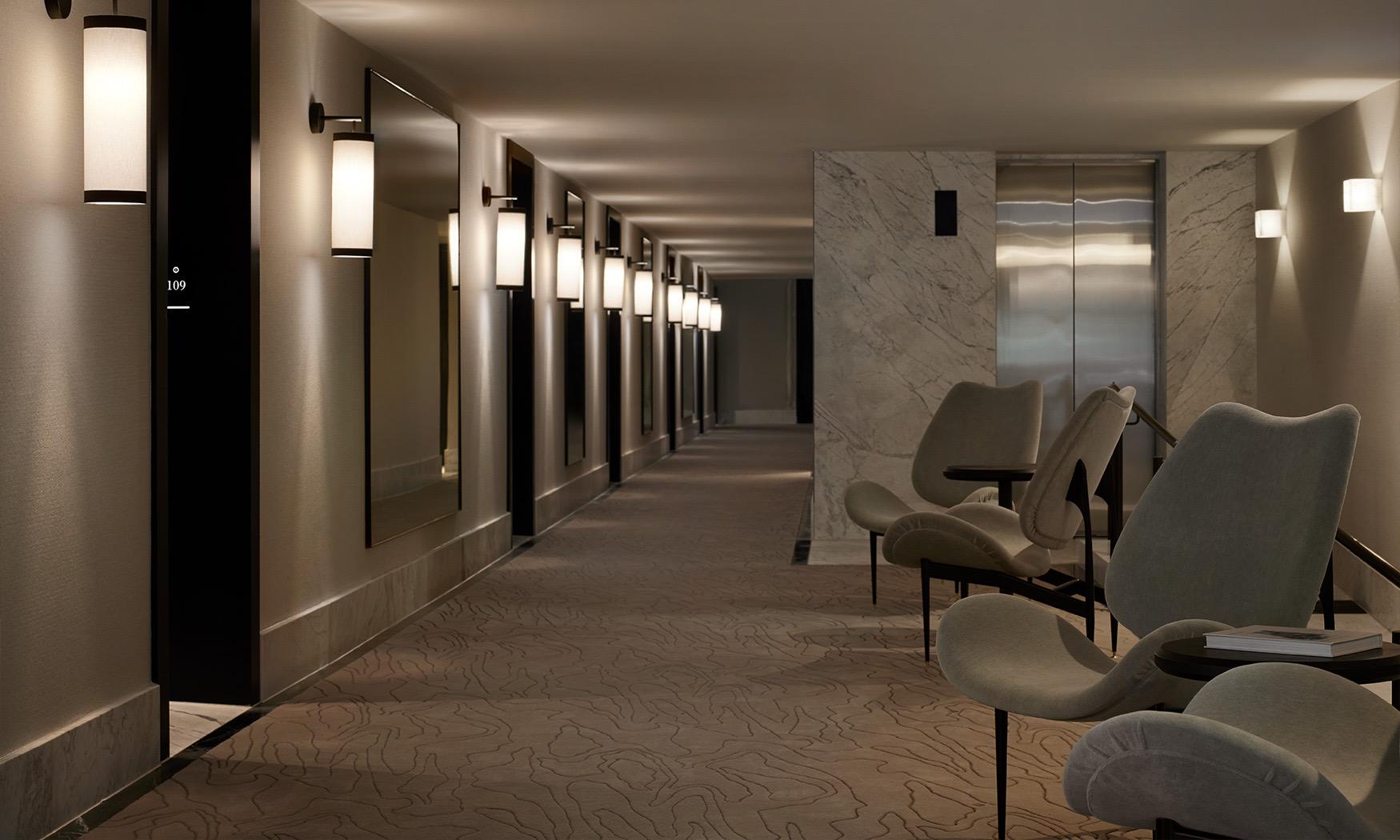 A long hotel corridor with wall sconces, framed mirrors, a marble lift lobby and upholstered chairs at the far end.