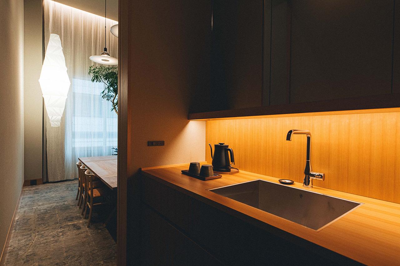 A wooden kitchenette counter with a sink, a black kettle and two cups, with a dining table and a paper pendant light visible through a doorway.