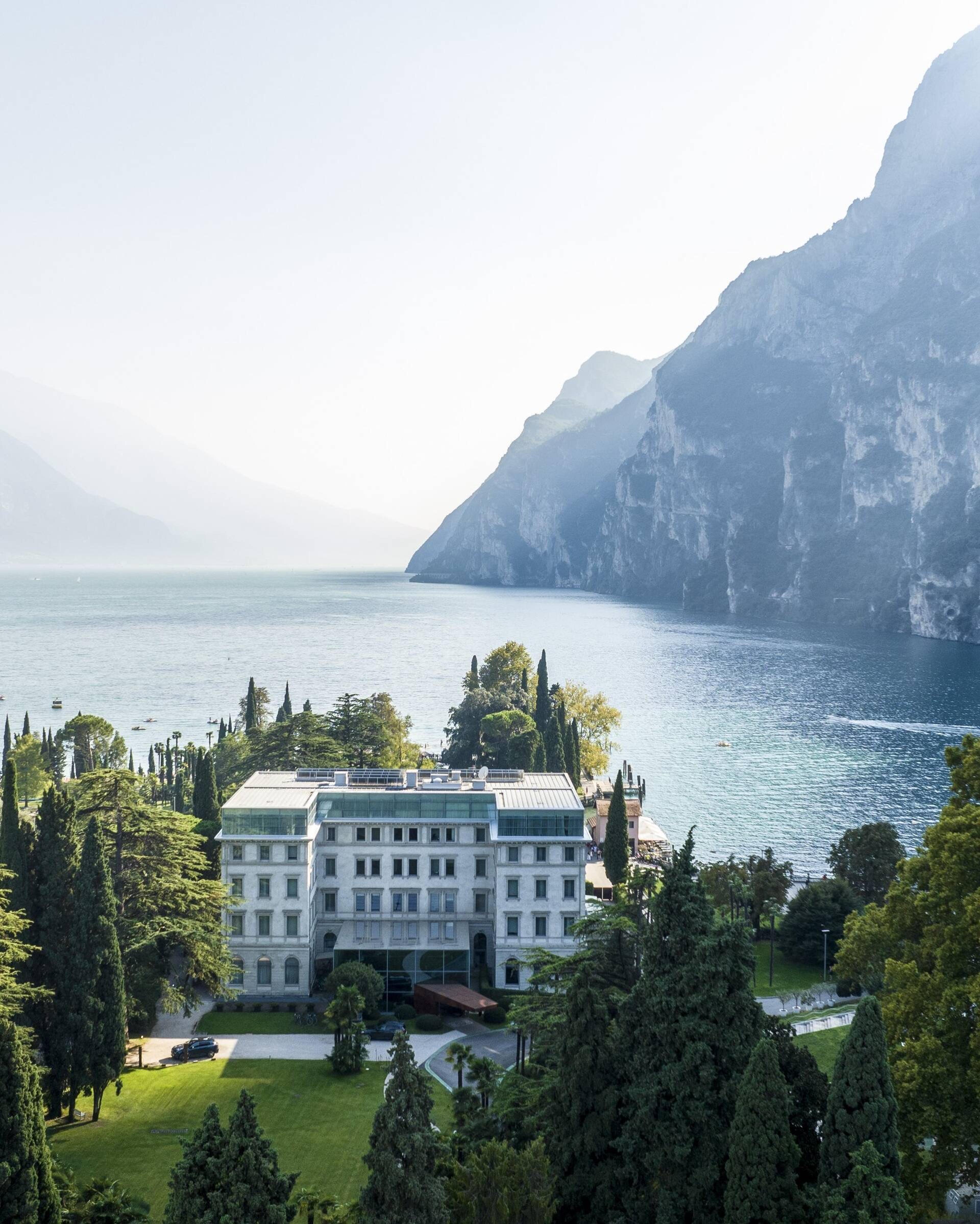 An aerial view of the hotel between pine trees and a lake, with mountains rising on the far shore.