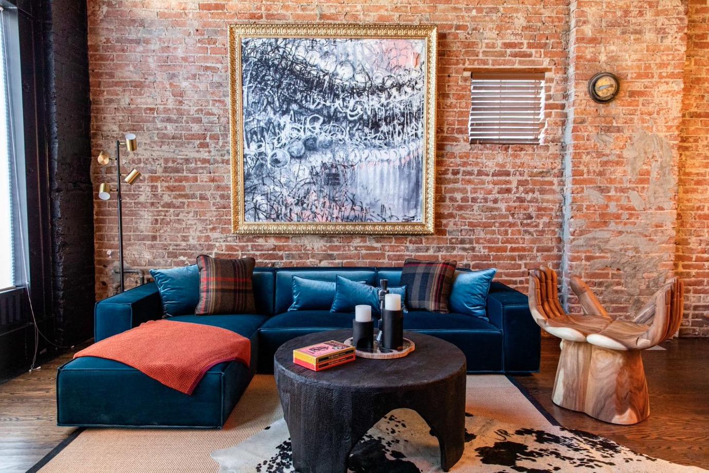 A living room with an exposed brick wall, a navy velvet corner sofa, a round black coffee table, a sculptural wood chair and a large framed abstract painting.