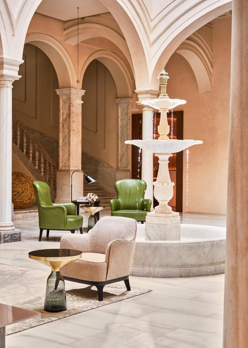 A tiered marble fountain stands under a vaulted arcade, flanked by green leather armchairs and a round brass side table, with a staircase visible behind.
