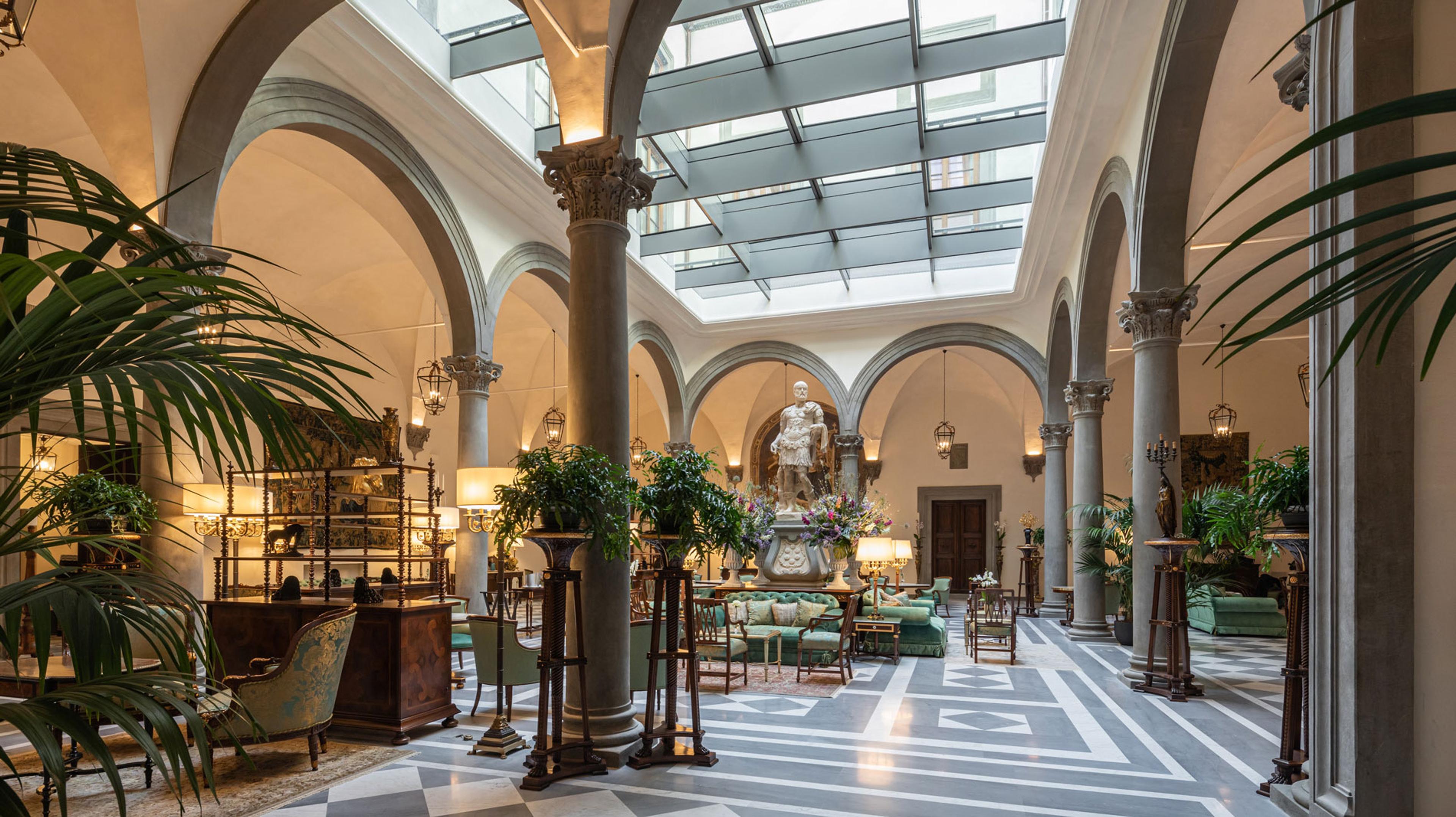a stone courtyard with a glass roof, arched colonnades on both sides, a classical statue among flowers, and green velvet sofas on a black and white marble floor