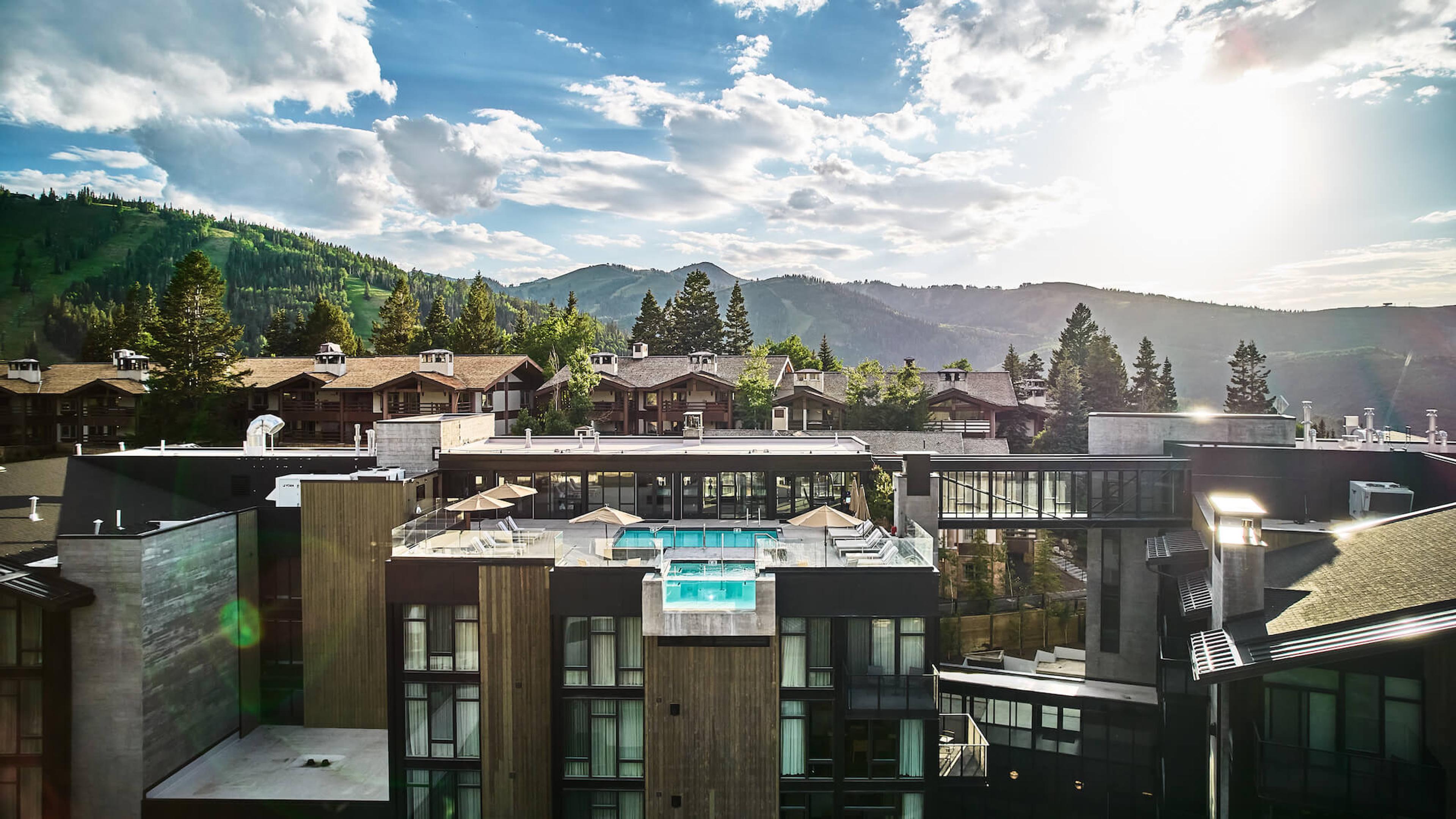 A rooftop pool and hot tub on a hotel terrace seen from above, with timber-clad buildings, pine trees and mountains behind under a partly cloudy sky