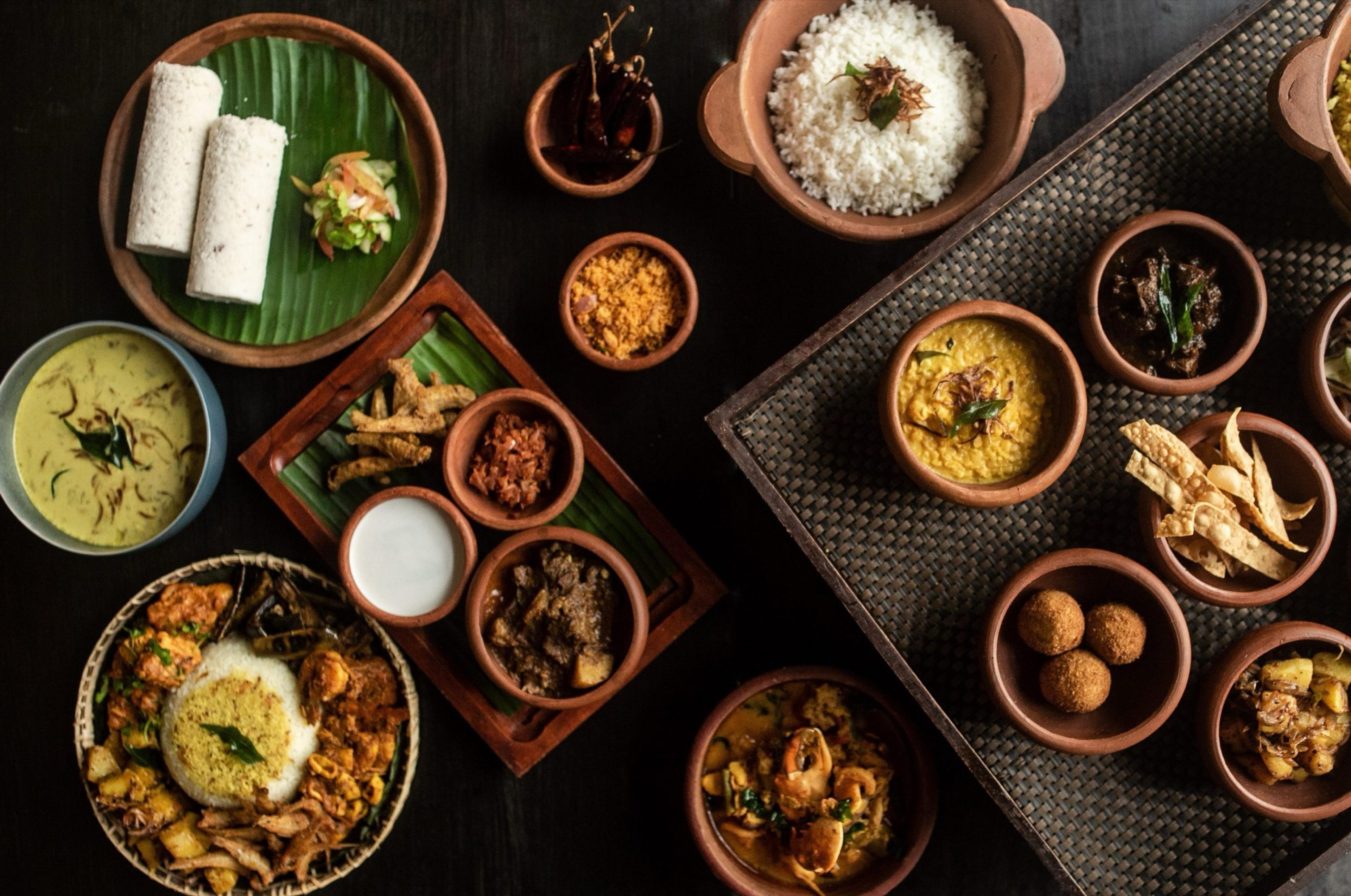 A spread of Sri Lankan dishes in small clay and ceramic bowls seen from above, including rice, curries, sambols and rolled pancakes on a banana leaf.