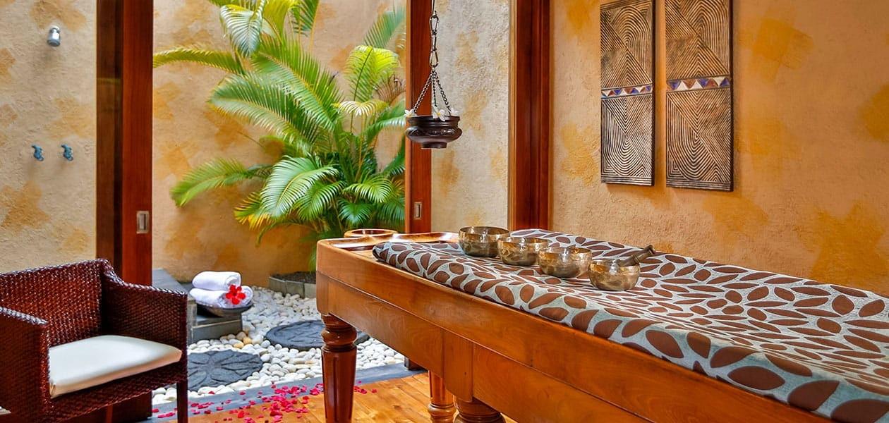 A massage table laid with small bowls in an open-air treatment room, with rose petals on the floor and a potted palm in the corner.