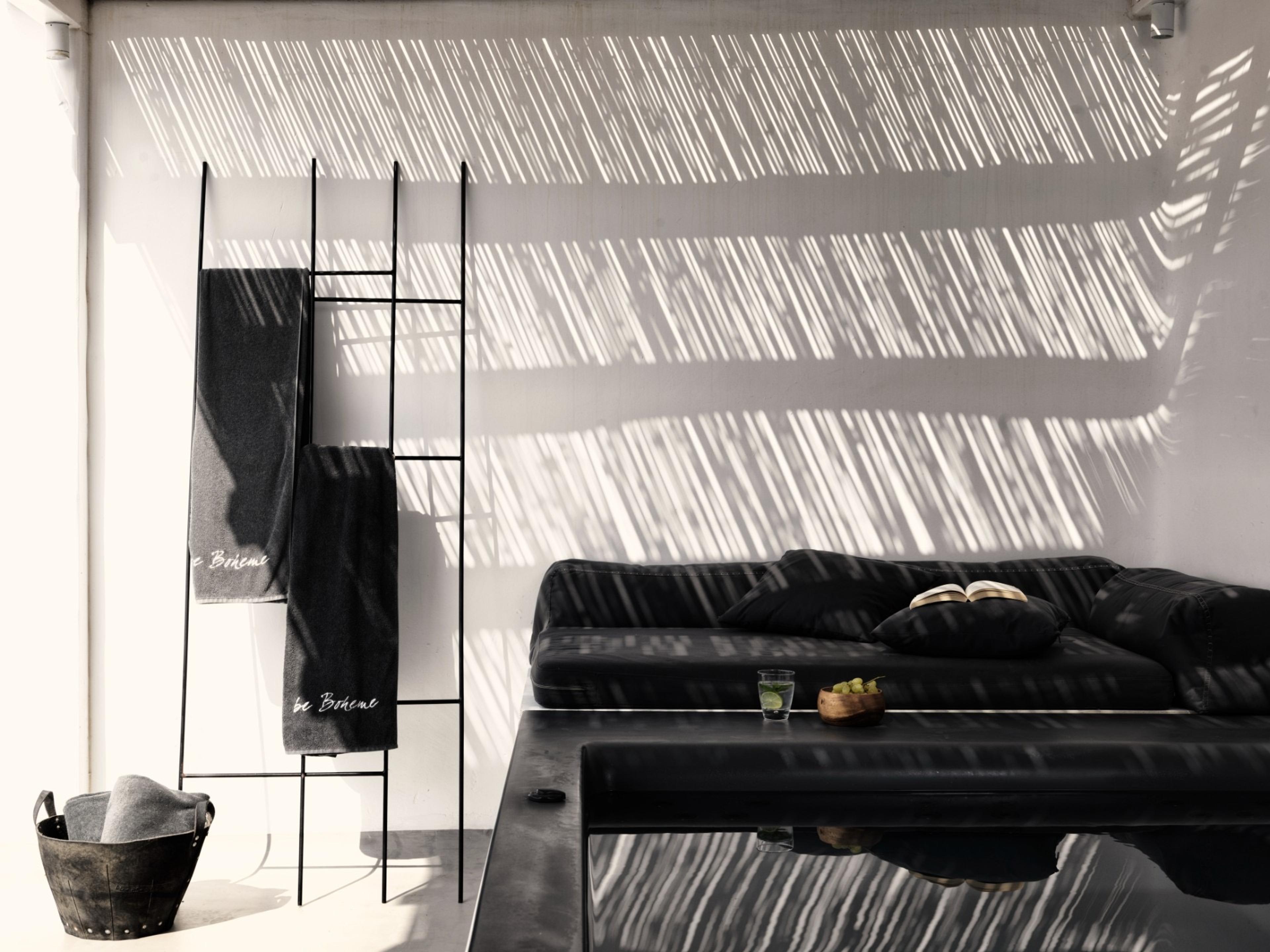 Grey towels marked be Boheme hang from a ladder rack beside a low black table and sofa, striped with afternoon shadow.