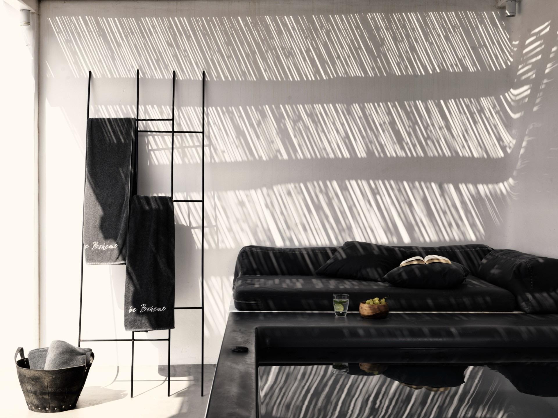 Grey towels marked be Boheme hang from a ladder rack beside a low black table and sofa, striped with afternoon shadow.