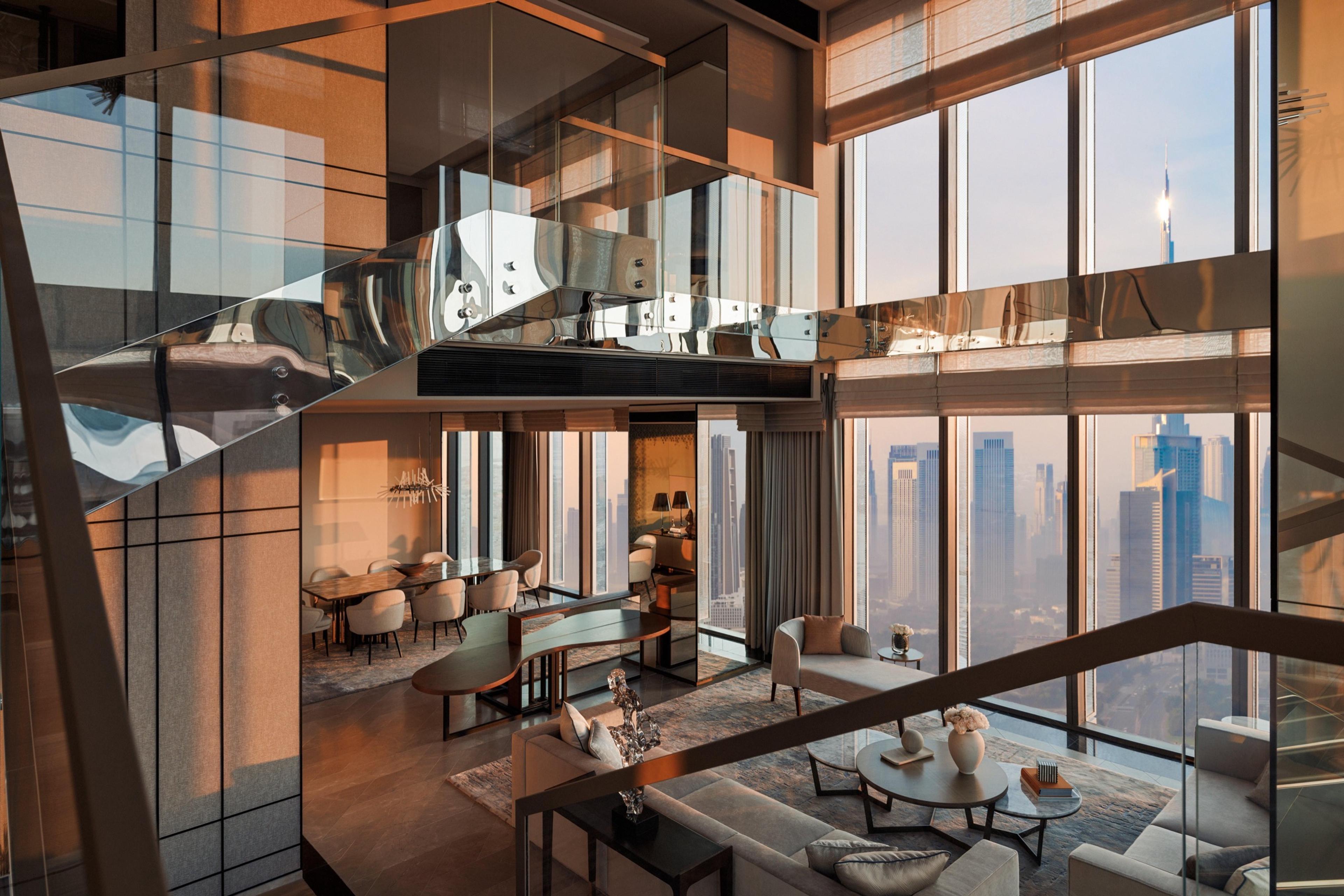 A two-storey suite seen from an upper landing, with a mirrored ceiling beam, a dining table below, a staircase, and floor-to-ceiling windows showing a sunset skyline.