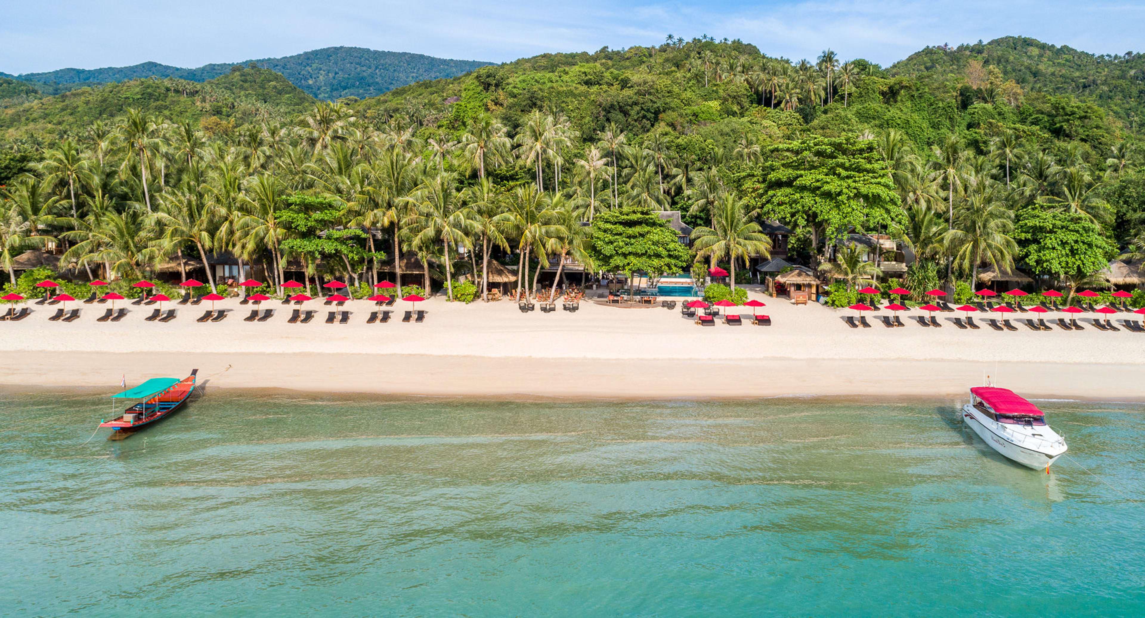 Red umbrellas and sun loungers line a white sand beach in front of palm trees and forested hills, with a longtail boat and a motorboat anchored in the turquoise water.