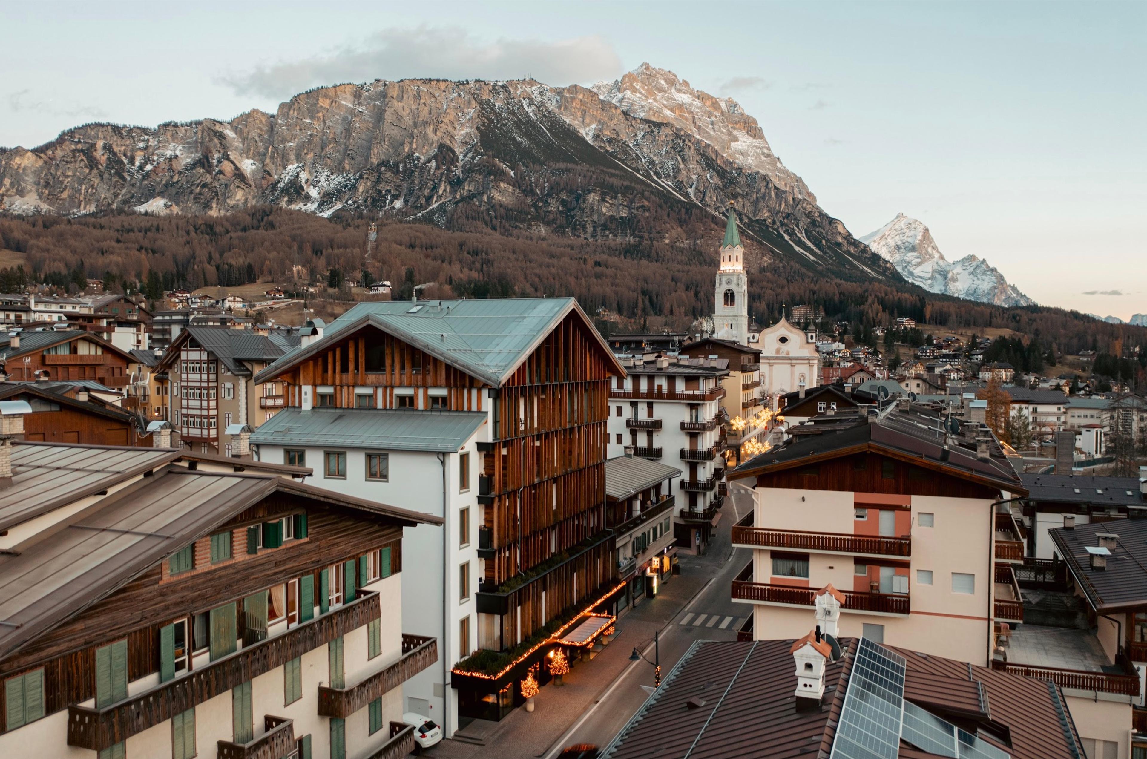 An elevated view over Cortina d'Ampezzo's rooftops and a church tower, with the hotel among them and jagged peaks behind.