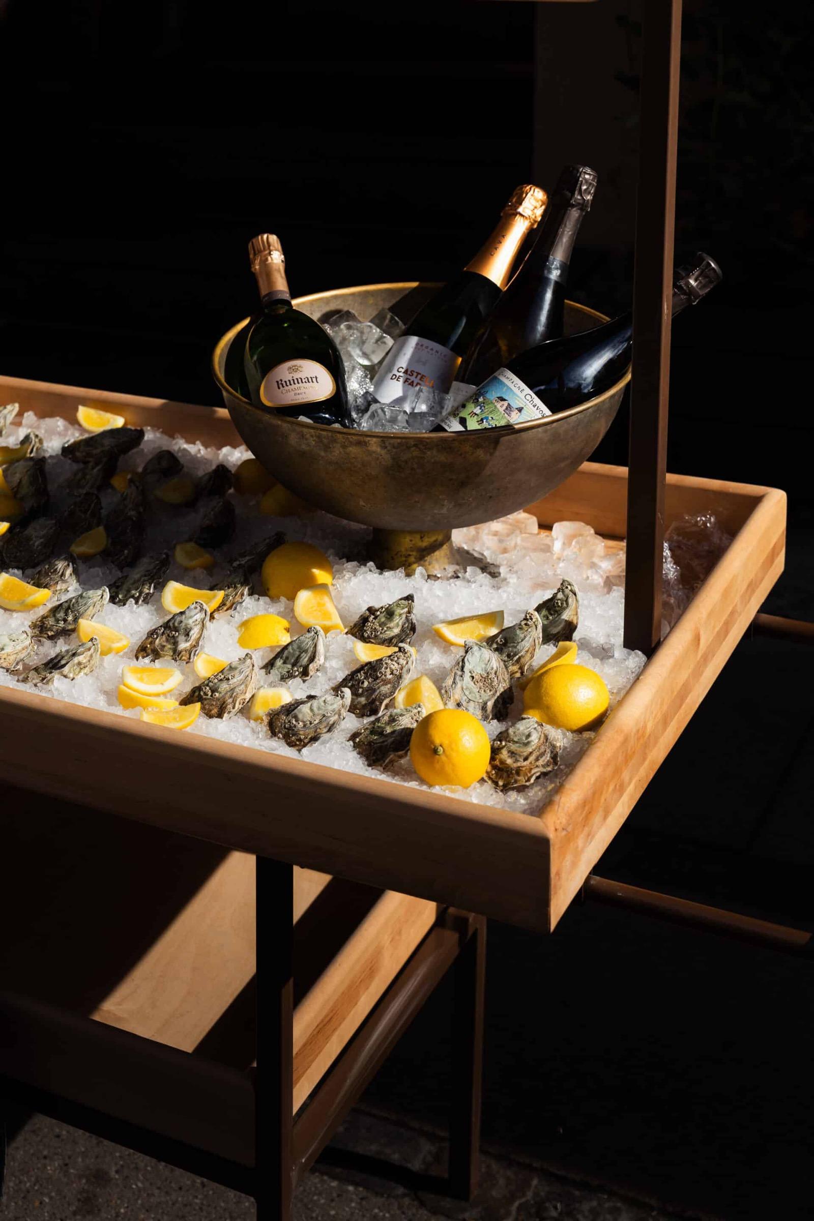 A wooden tray of oysters on ice with lemon wedges, and three champagne bottles chilling in a brass bowl, lit against a dark background.
