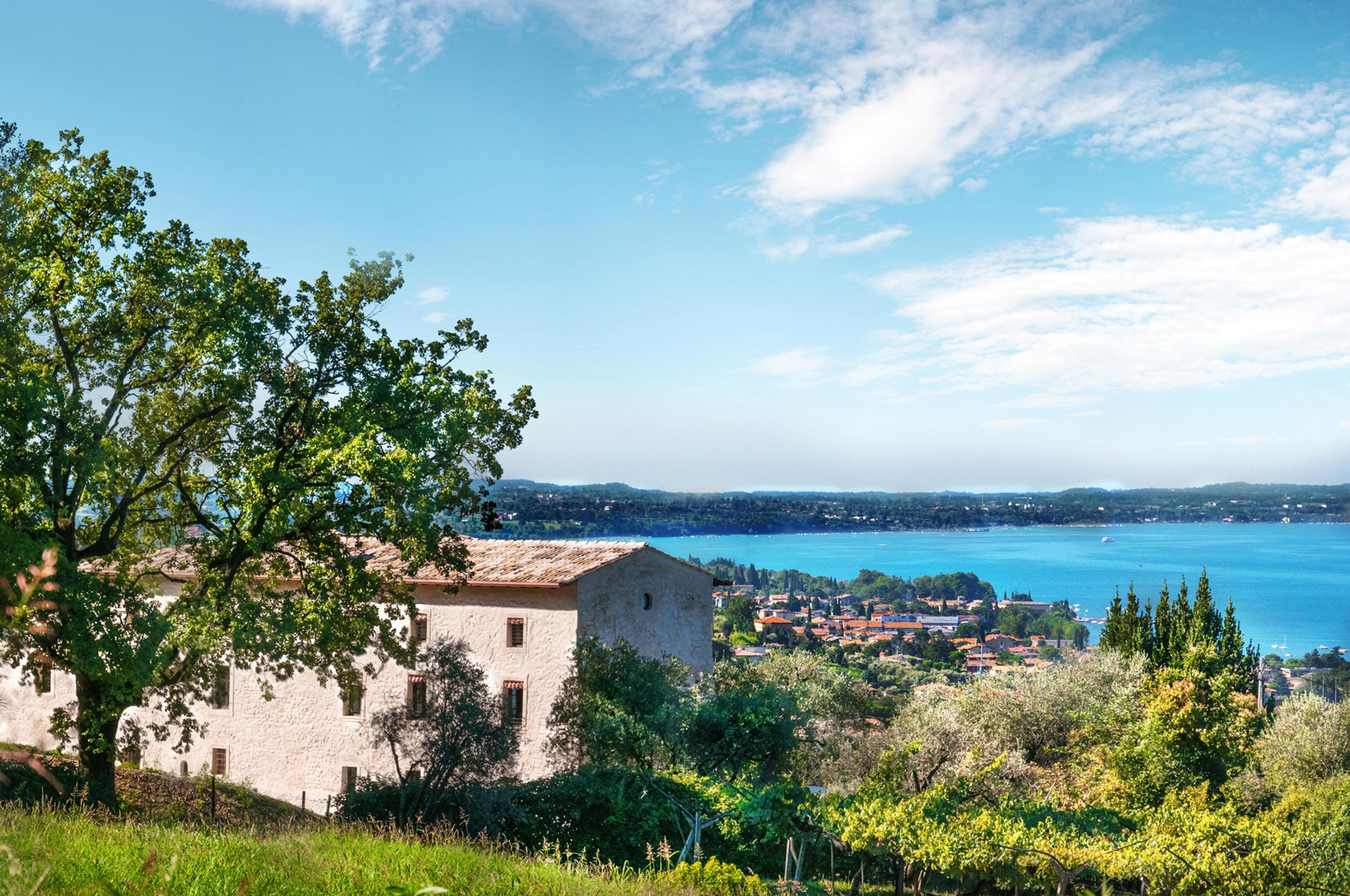 A stone villa with a tiled roof on a hillside, framed by a large tree, overlooking a lakeside town and a wide blue lake.