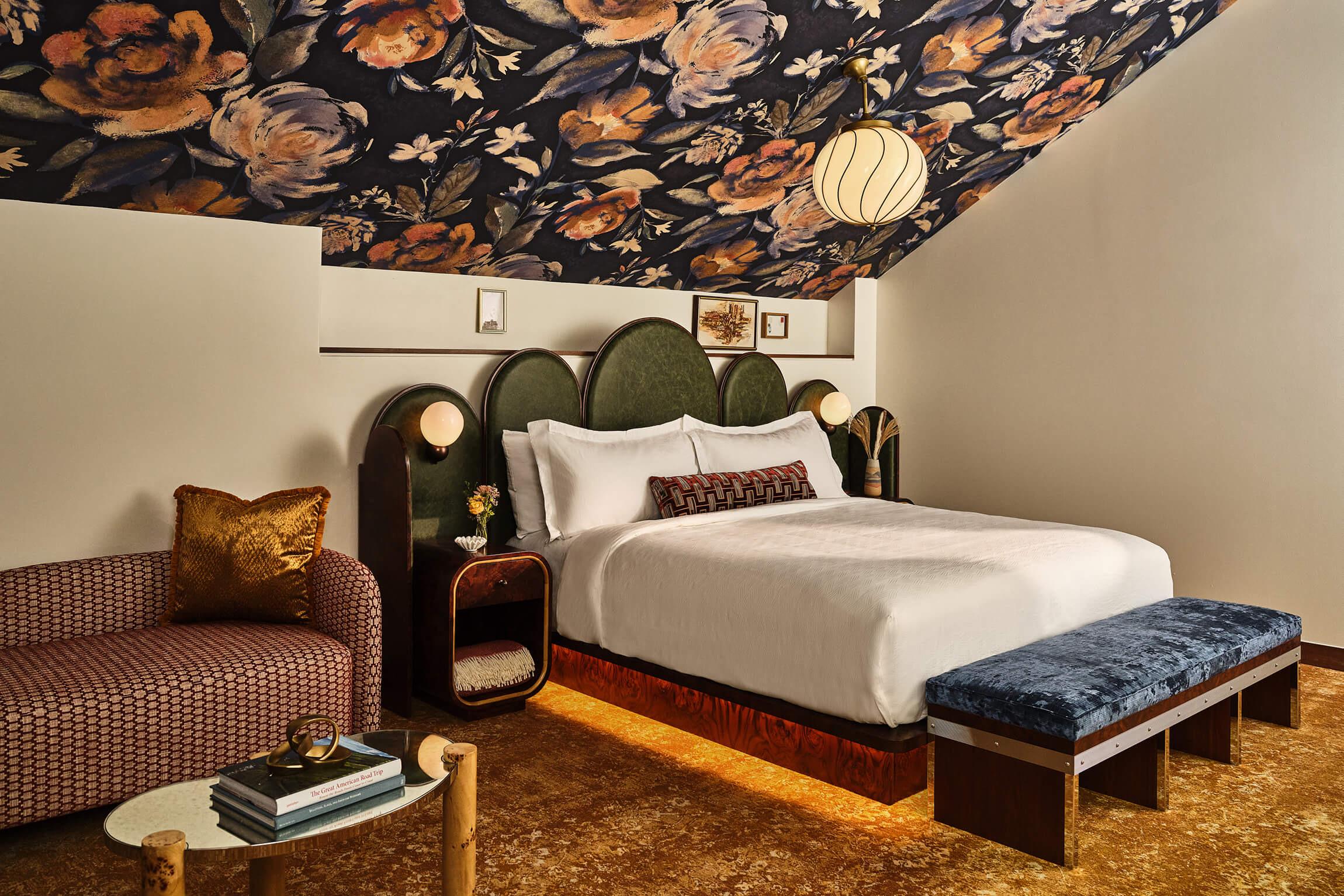 A bedroom under a sloped ceiling covered in dark floral wallpaper, with a white bed, a green arched headboard and a blue upholstered bench at its foot.