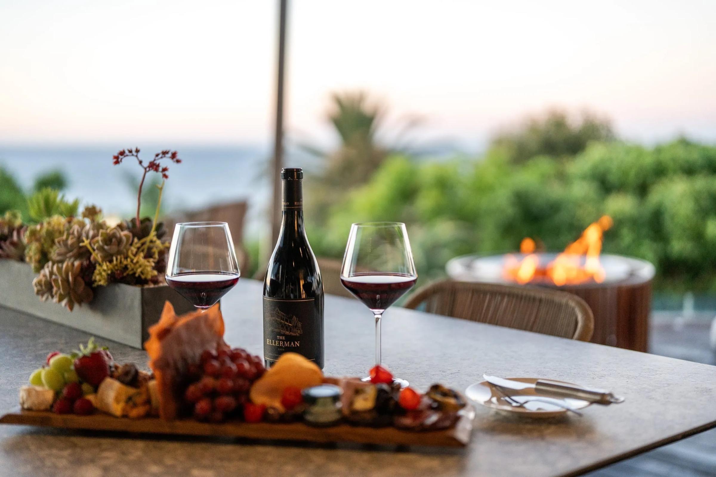 Two glasses of red wine and a bottle labelled The Ellerman stand on a table beside a platter of cheese, cured meat and fruit, with a lit fire bowl and trees beyond.