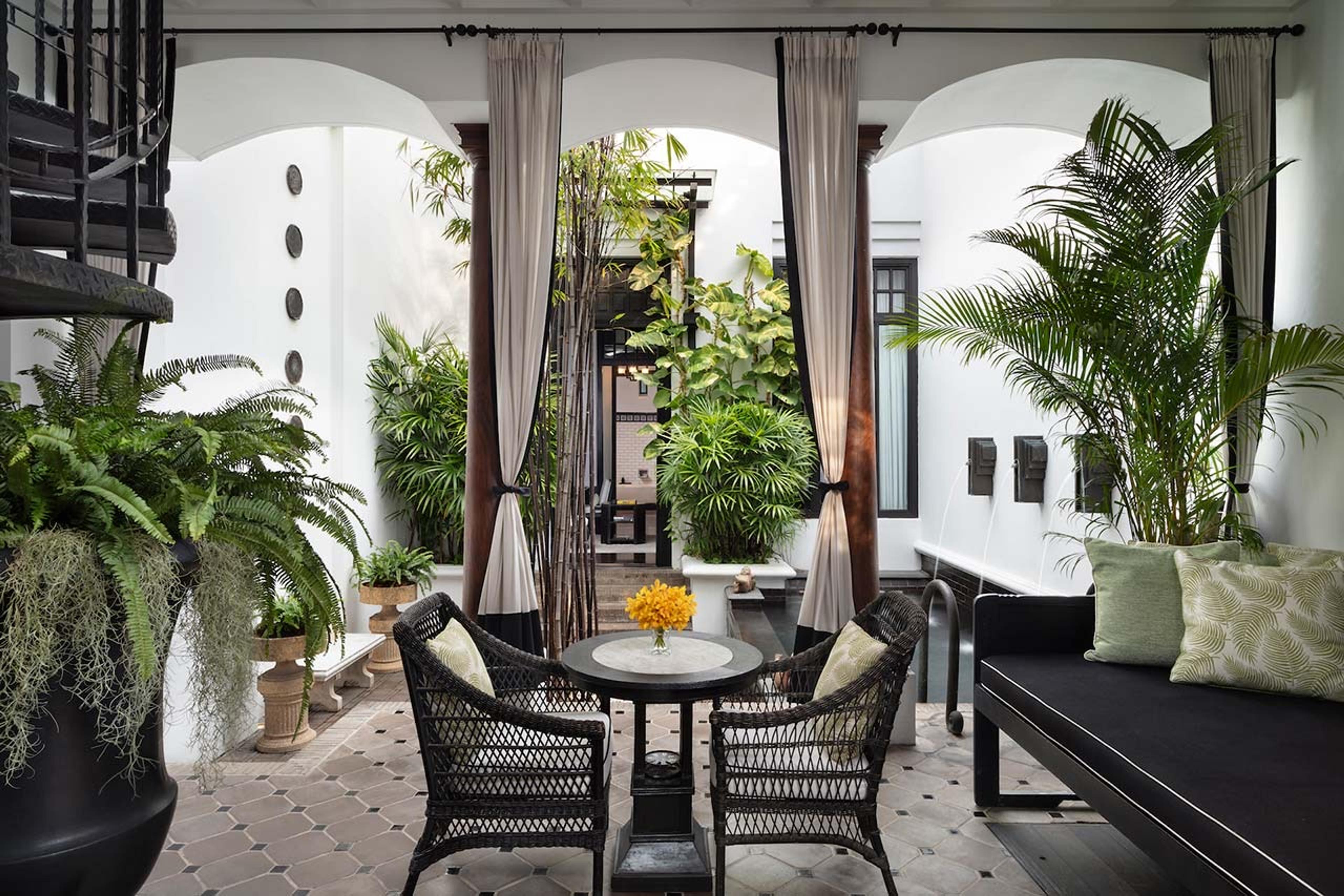 A black wrought-iron spiral staircase and potted ferns and palms surround a small wicker table and chairs in a courtyard, curtained archways and a trellis roof overhead.