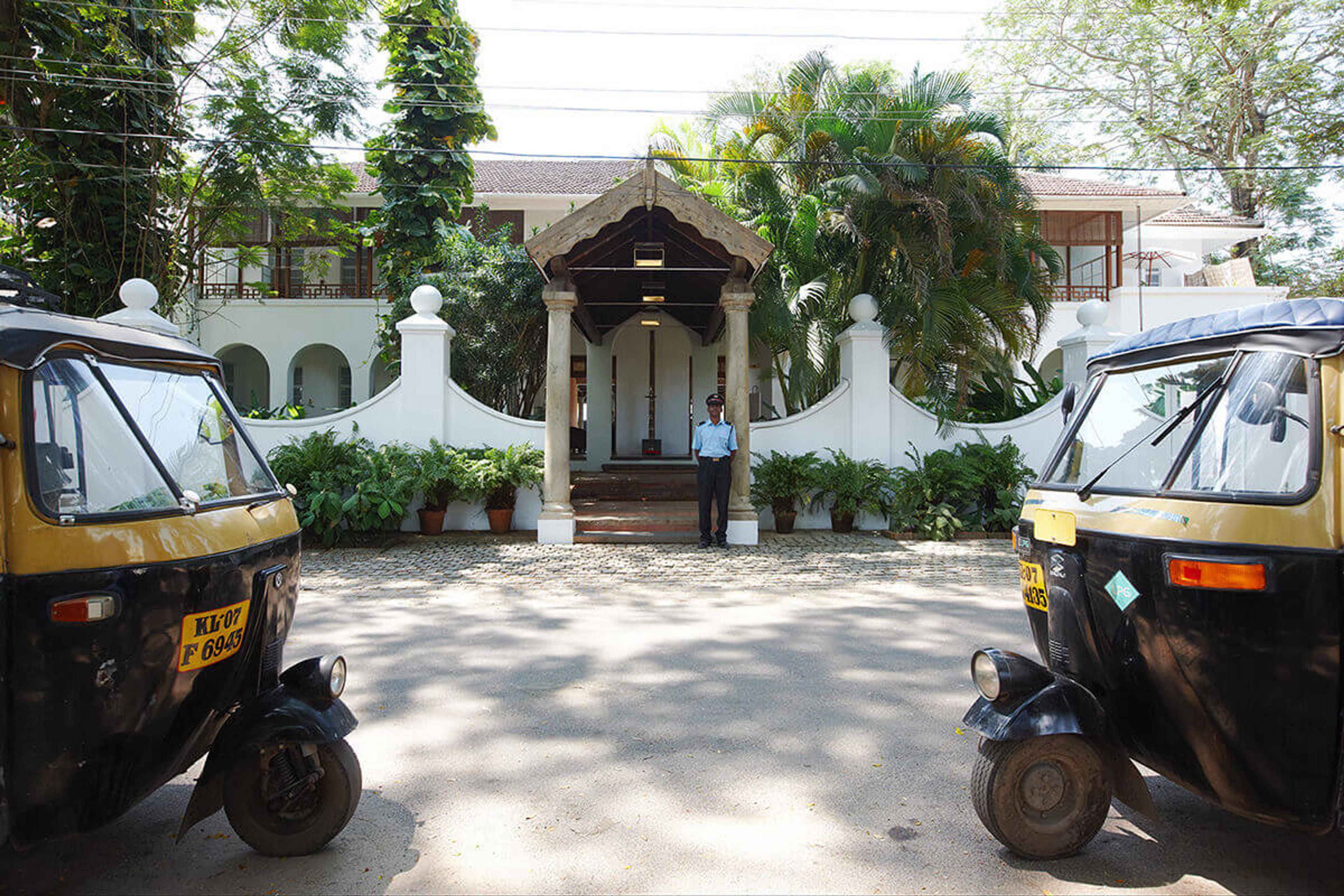 A white colonial-style building entrance framed by two parked auto-rickshaws, with a uniformed man standing at the door.