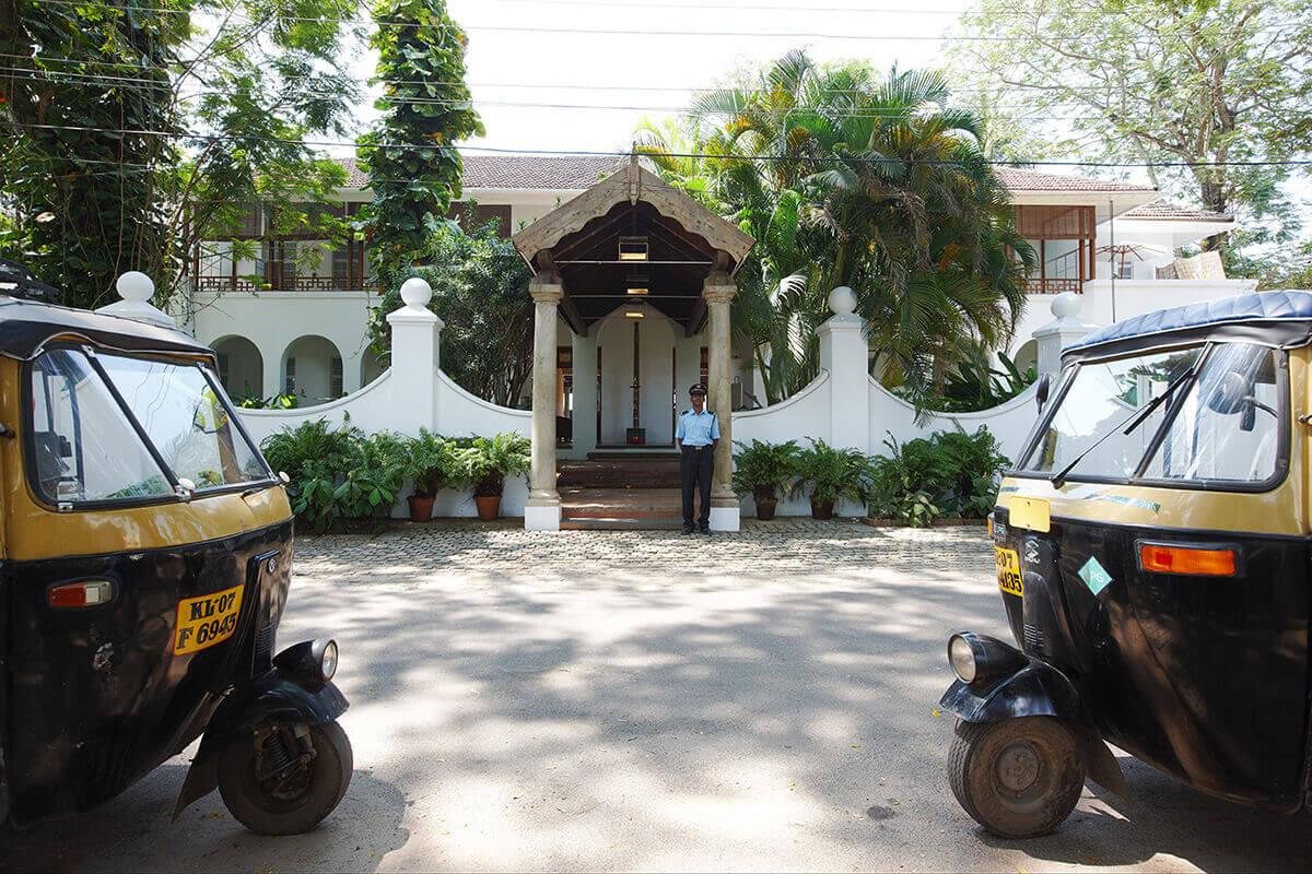 A white colonial-style building entrance framed by two parked auto-rickshaws, with a uniformed man standing at the door.