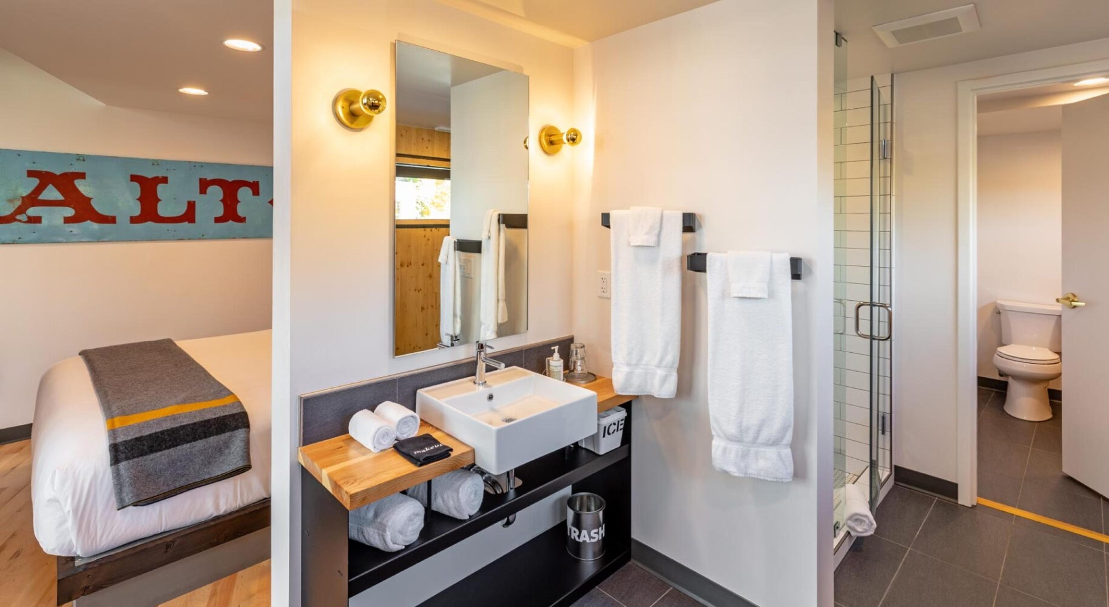 A bathroom with a rectangular white basin on a dark wood and steel vanity, folded white towels, brass wall lights, and a glass-walled shower with white tiles.