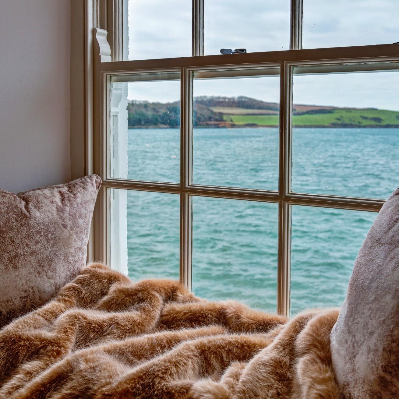 A window seat heaped with a faux fur throw and velvet cushions, looking out through a sash window over an estuary to green hills opposite.