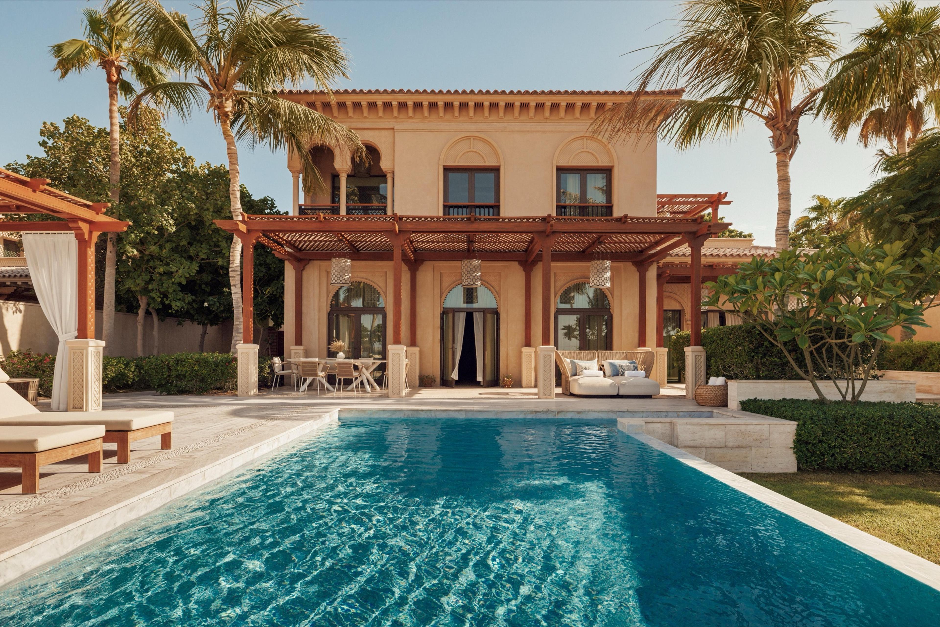 A two-story sand-colored villa with arched windows and a wood pergola stands behind an L-shaped pool lined with loungers and palm trees.