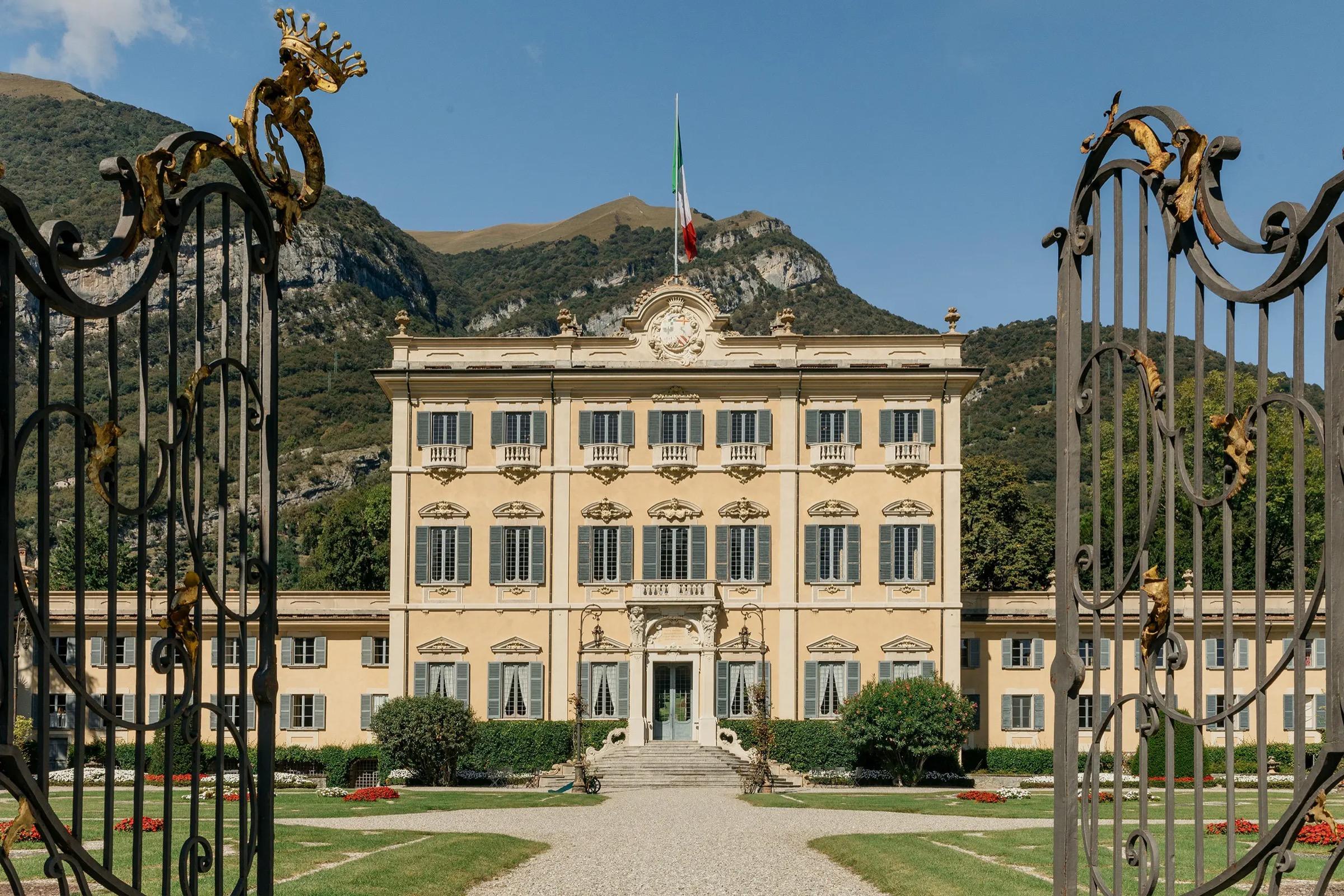 A cream-colored villa with shuttered windows and an Italian flag stands beyond a wrought iron gate, framed by gardens and mountains.