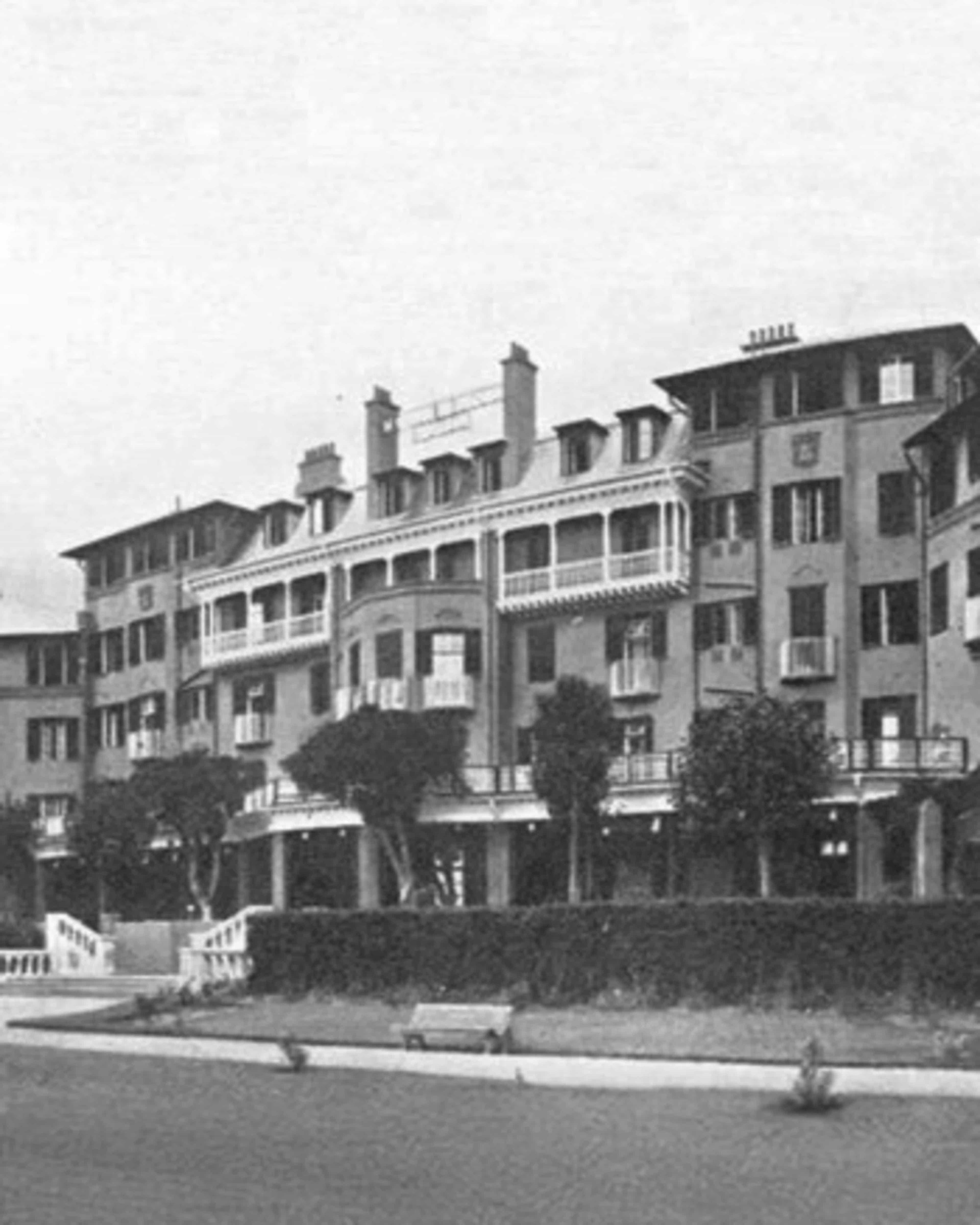A black and white photograph of a multi-storey building with balconies, dormer windows and chimneys, behind a hedge and a lawn.