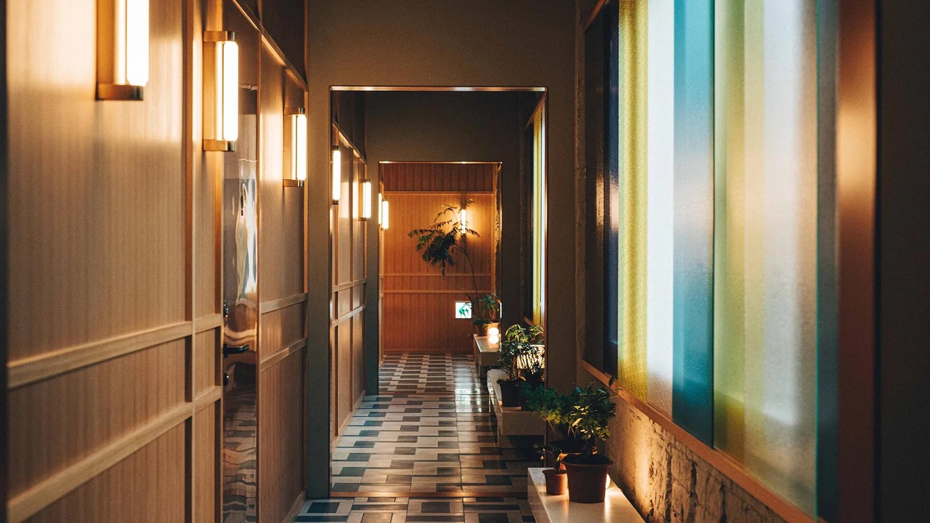A hotel corridor at night with wall lamps, a checkerboard tile floor, potted plants, and panels of coloured glass along one wall.