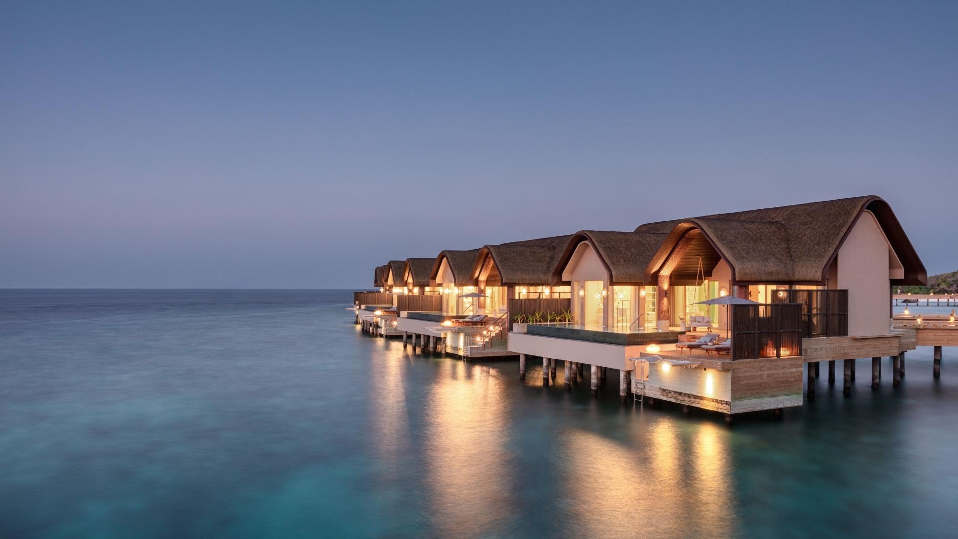 A row of over-water villas lit from within at dusk across still water.