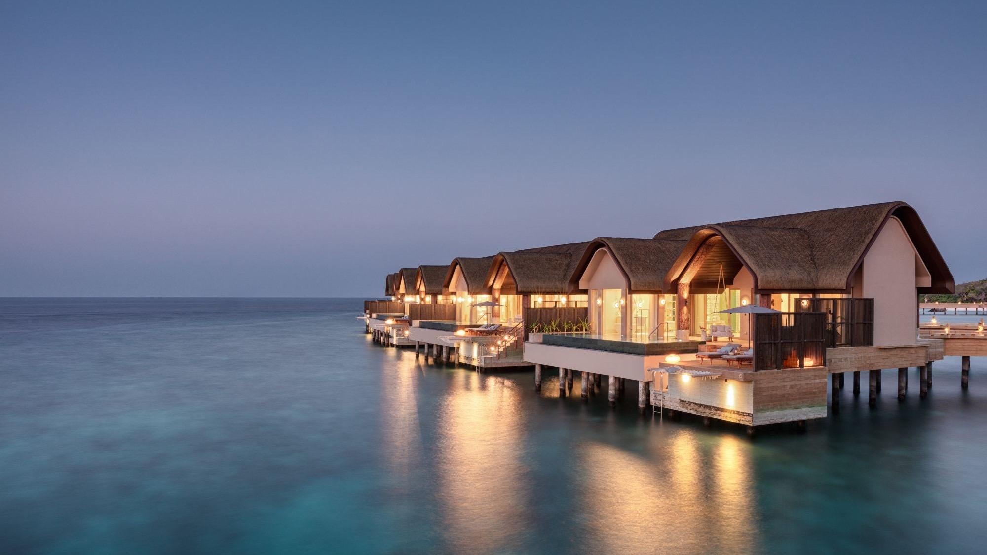 A row of over-water villas lit from within at dusk across still water.