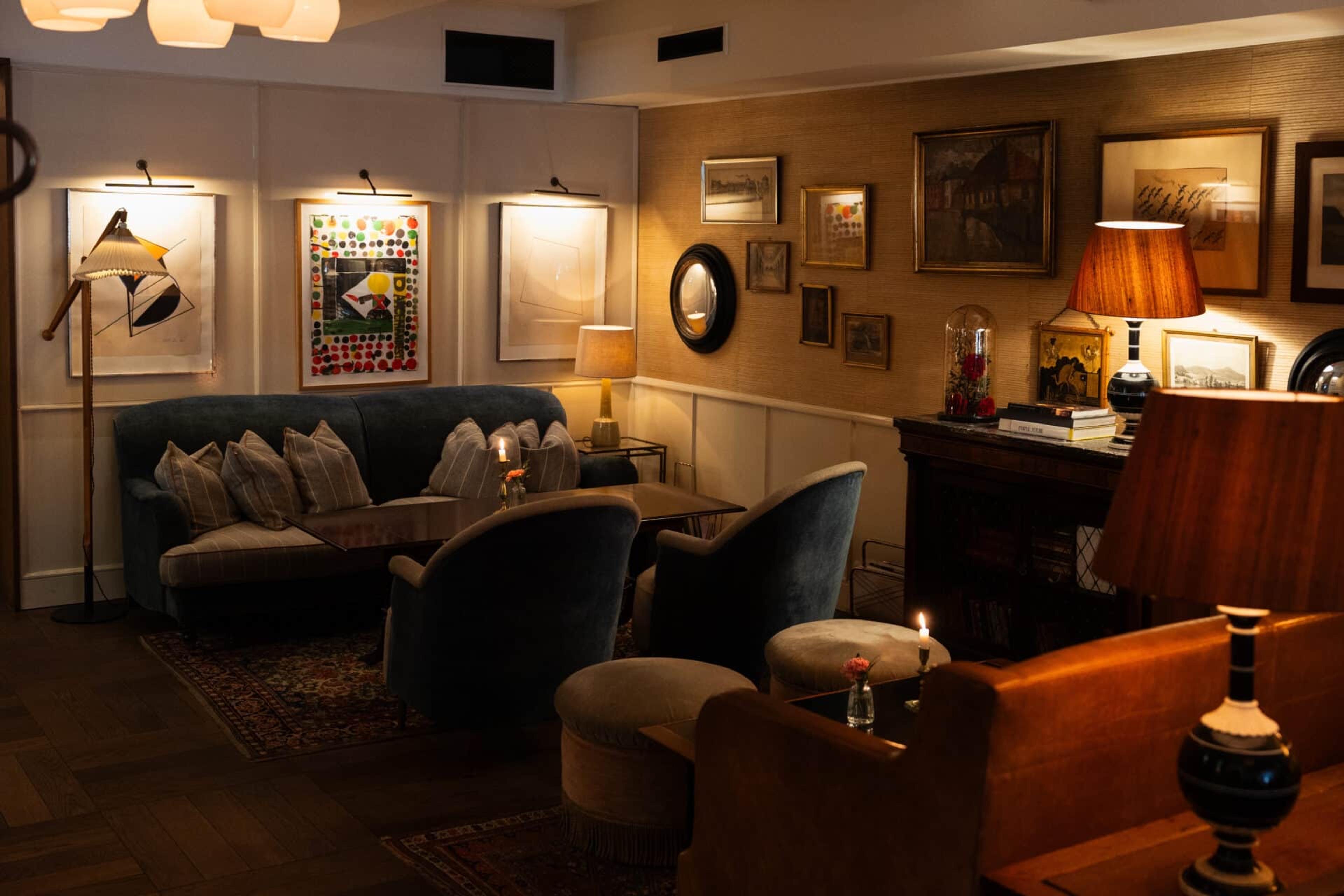 A dim lounge with two dark green armchairs and a matching sofa around a low table, framed art lit from above on cream walls, and lamps lit on a side cabinet.
