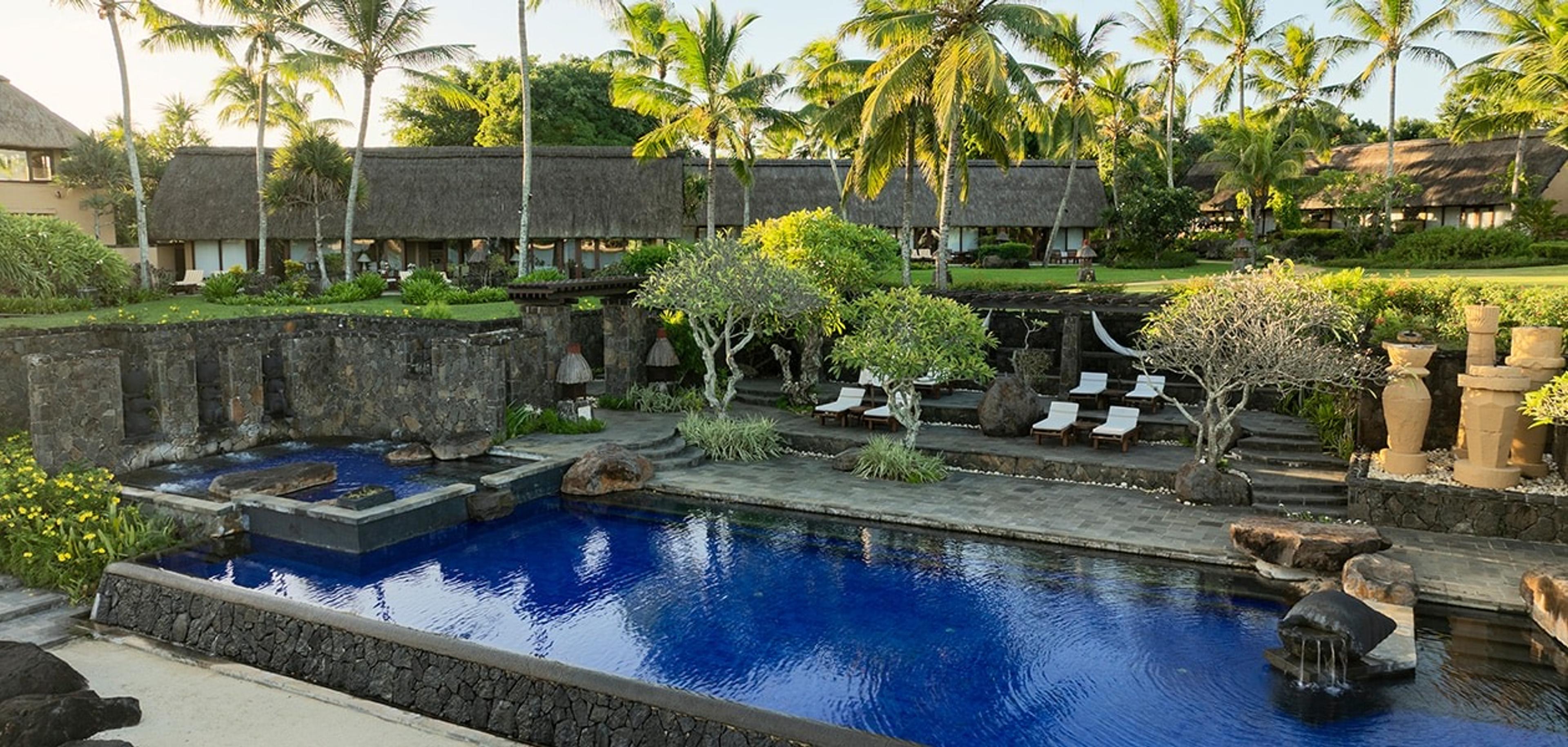 A stone-walled lap pool with a fountain jet, bordered by sun loungers and palm trees, with thatched buildings behind it at dusk.