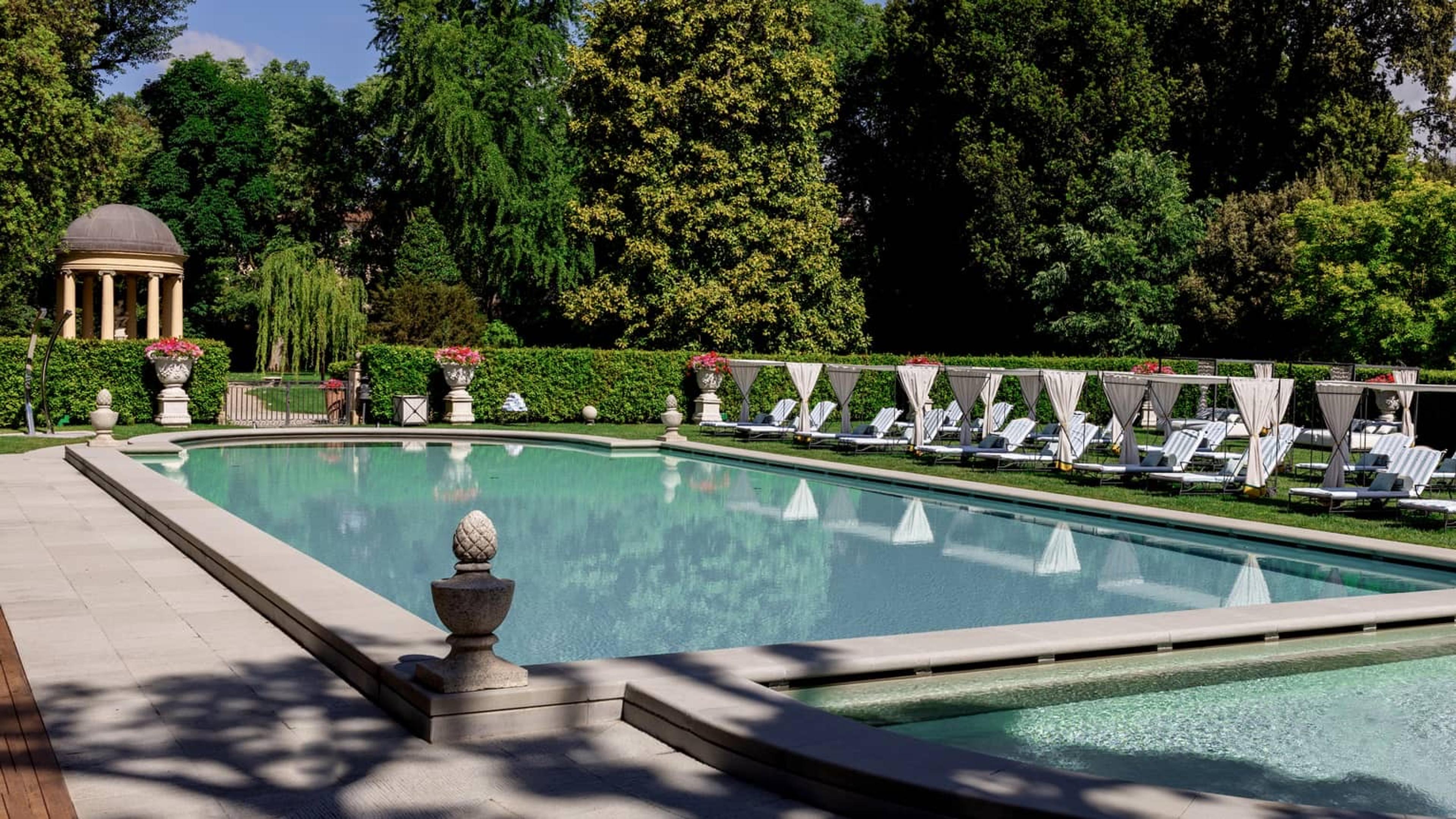 A rectangular swimming pool with a row of white lounge chairs and cabanas along one side, trees behind and a domed garden folly in the distance.