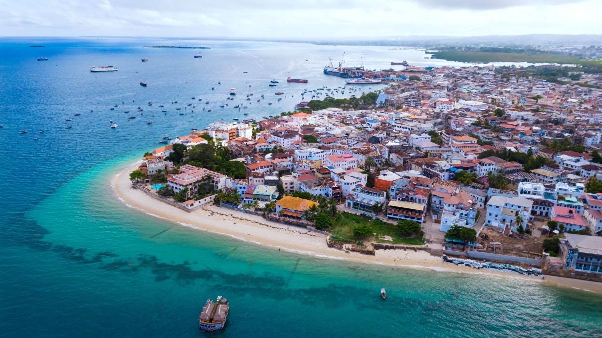 an aerial view of Stone Town's peninsula, its rooftops packed along a white sand beach against turquoise water with boats at anchor