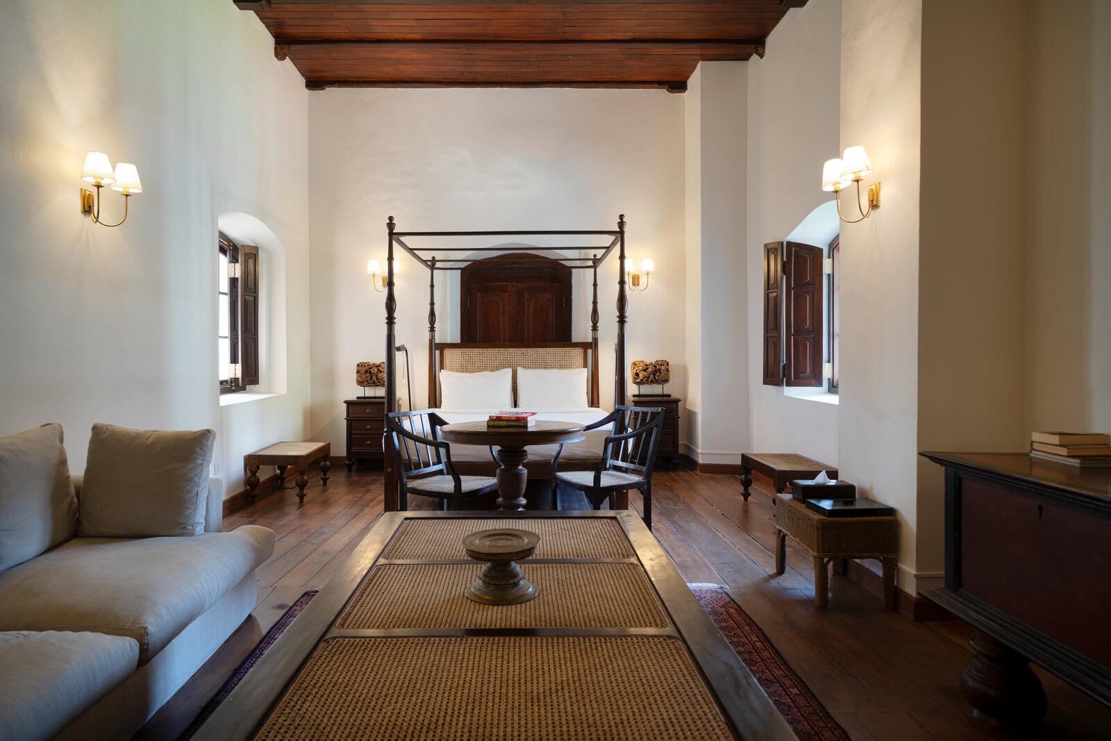 A vaulted room with a canopy bed in an arched alcove at the back, a round table and two chairs in the foreground, and wall sconces either side.