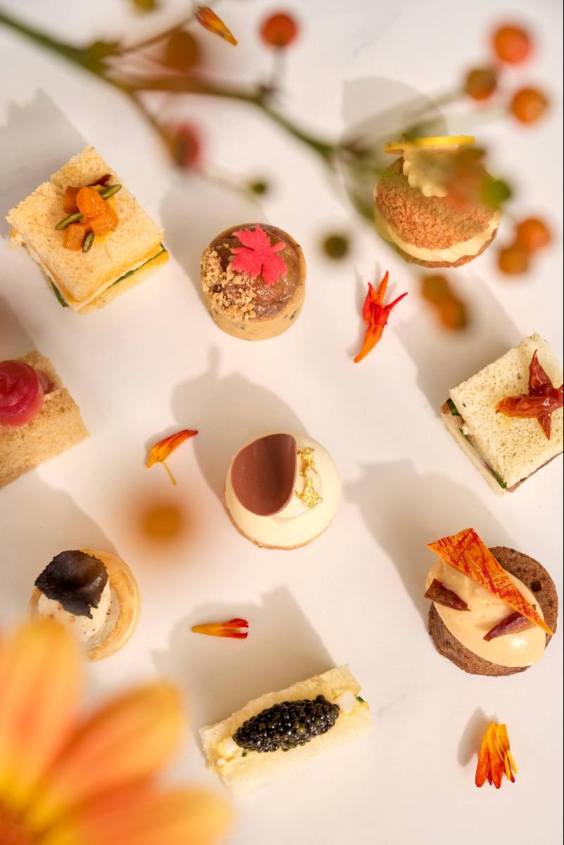 an overhead view of afternoon tea bites including a caviar finger sandwich, a chocolate dome and petits fours scattered with small orange flowers