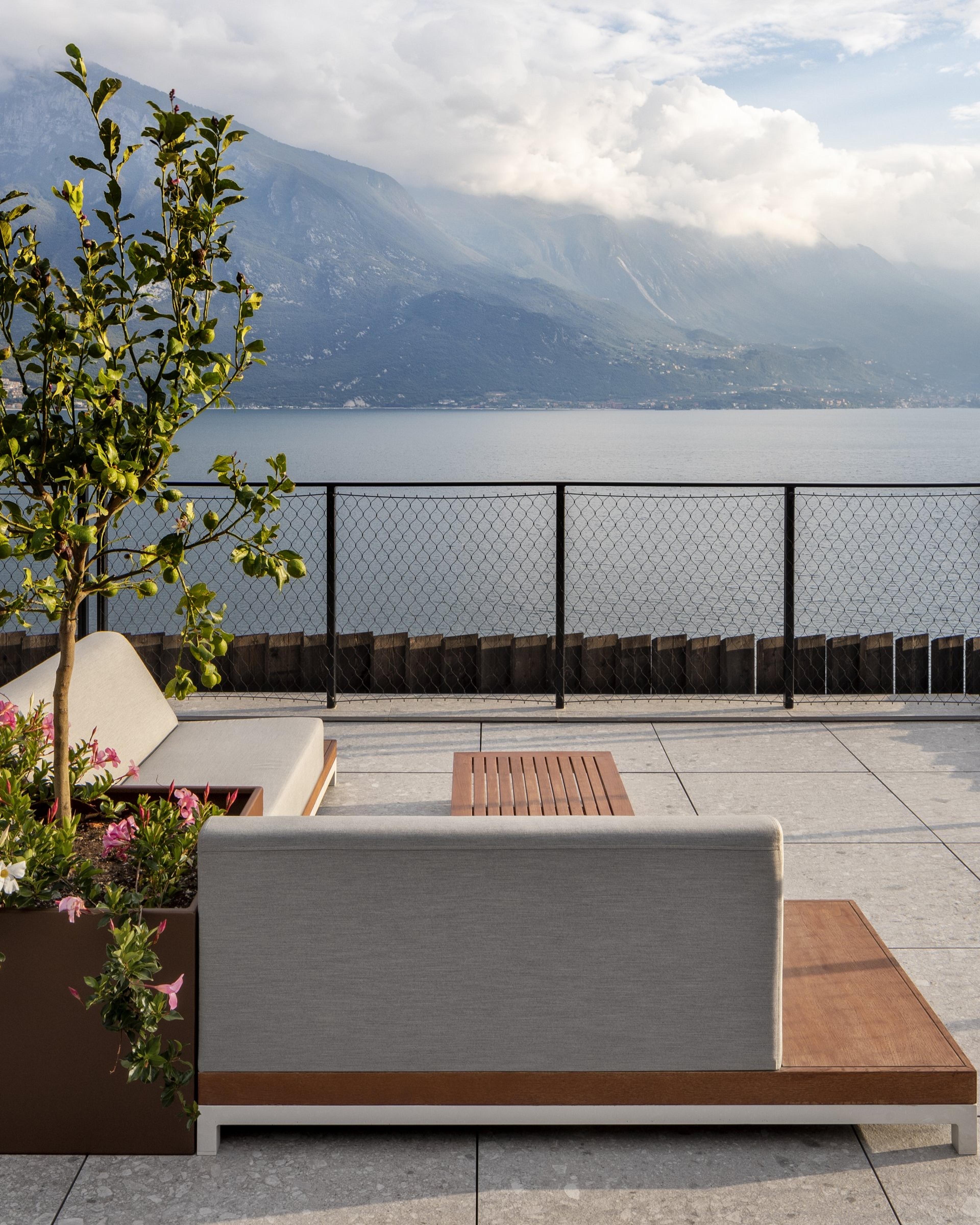 an outdoor terrace seating area with a lemon tree in a planter, overlooking a lake and mountains behind a wire fence