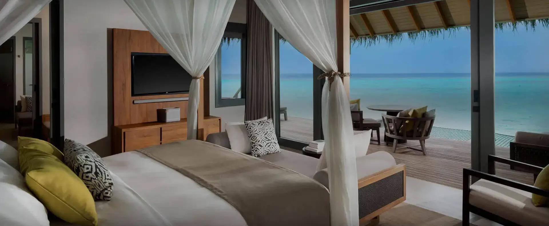 a bedroom with a curtained four-poster bed and a wooden console with a television, opening onto a covered deck with a dining table and chairs over the water