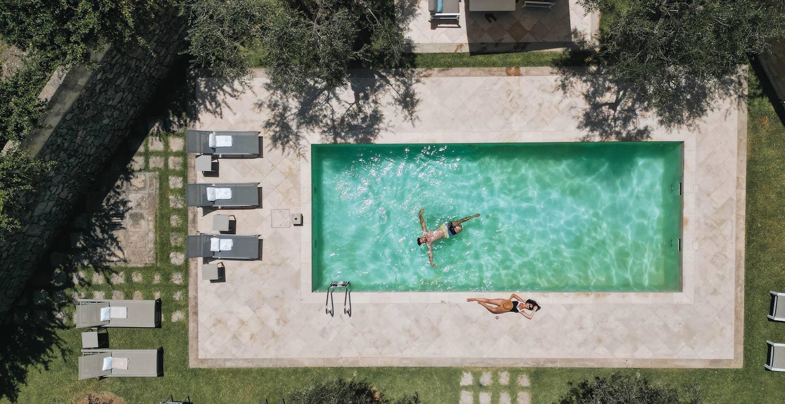 A swimming pool seen from directly overhead, with a paved terrace, sun loungers under trees, and two people in the water.