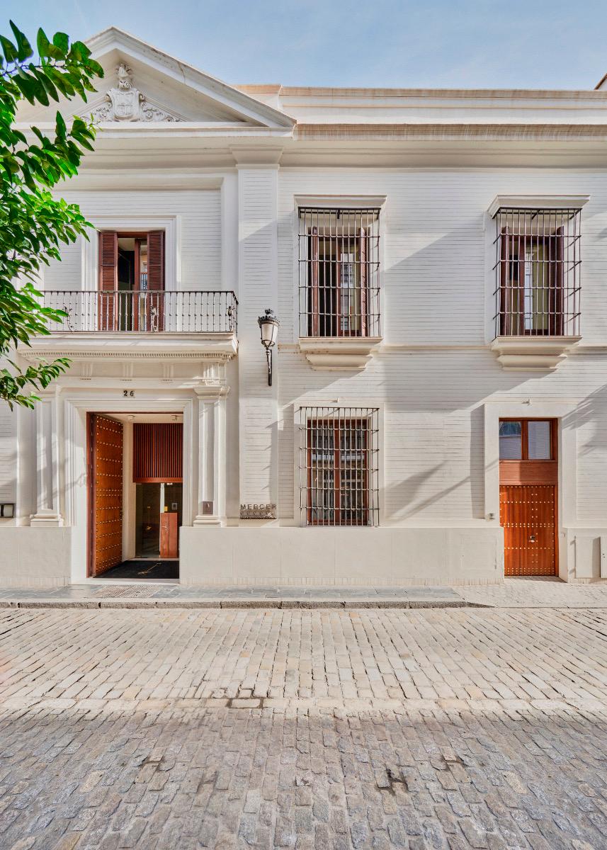 The hotel's pale stone facade rises over a cobbled street, with wrought iron balconies, barred windows, and a wooden door open at street level.