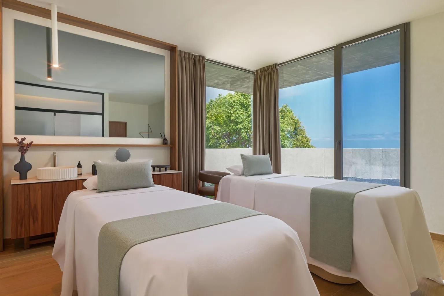 A spa treatment room with two covered massage beds, a long mirror, and a window looking onto trees and sky.