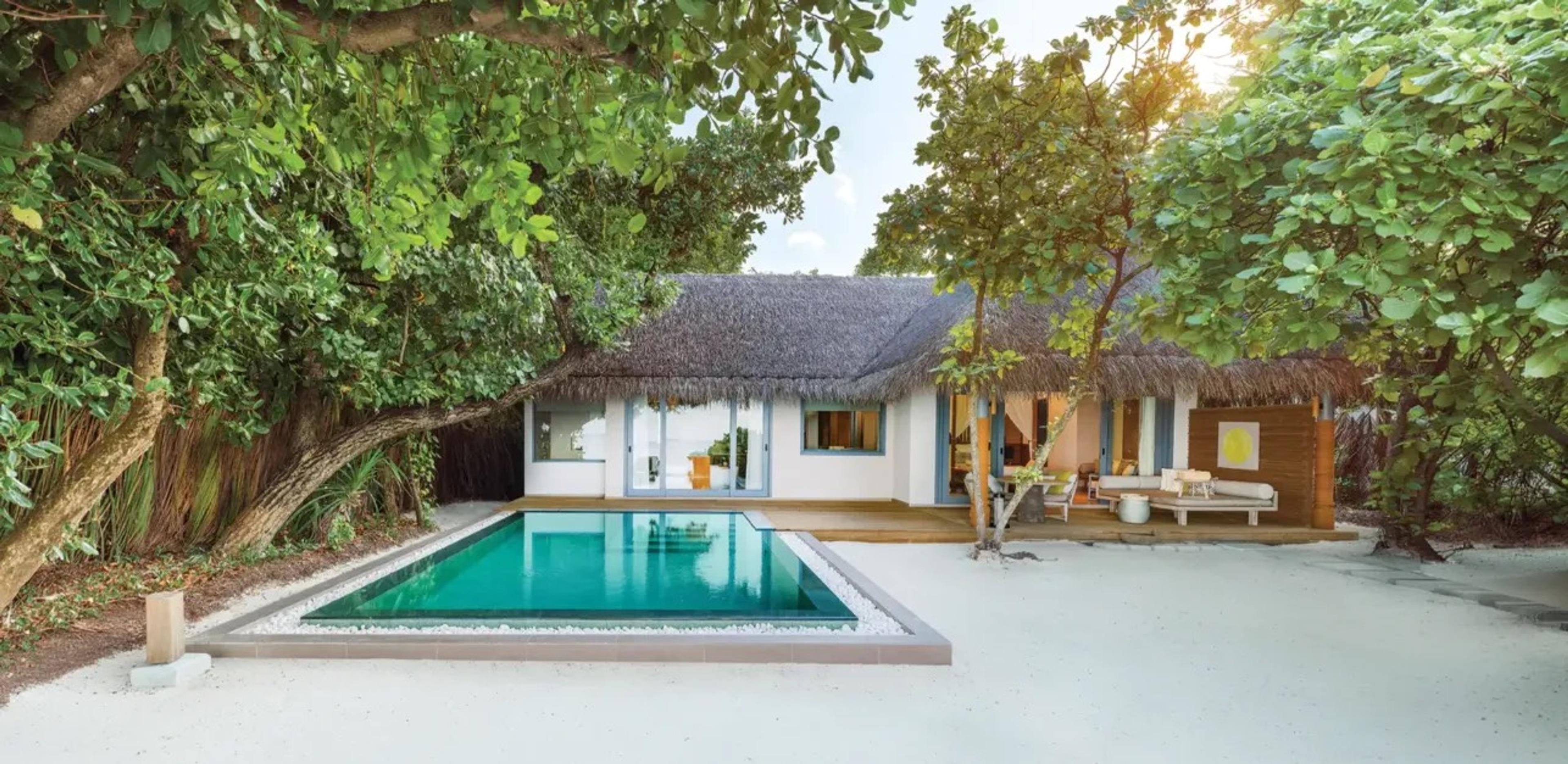 a white villa with blue-trimmed windows and a thatched roof, a rectangular pool on its sand terrace, framed by trees