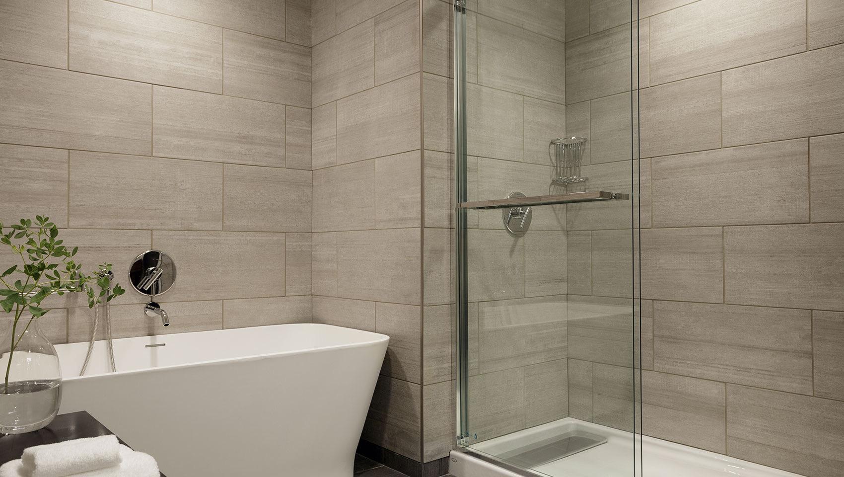 A freestanding white soaking tub beside a glass-walled shower, with tiled walls, a vase of greenery and folded towels on the tub's edge.