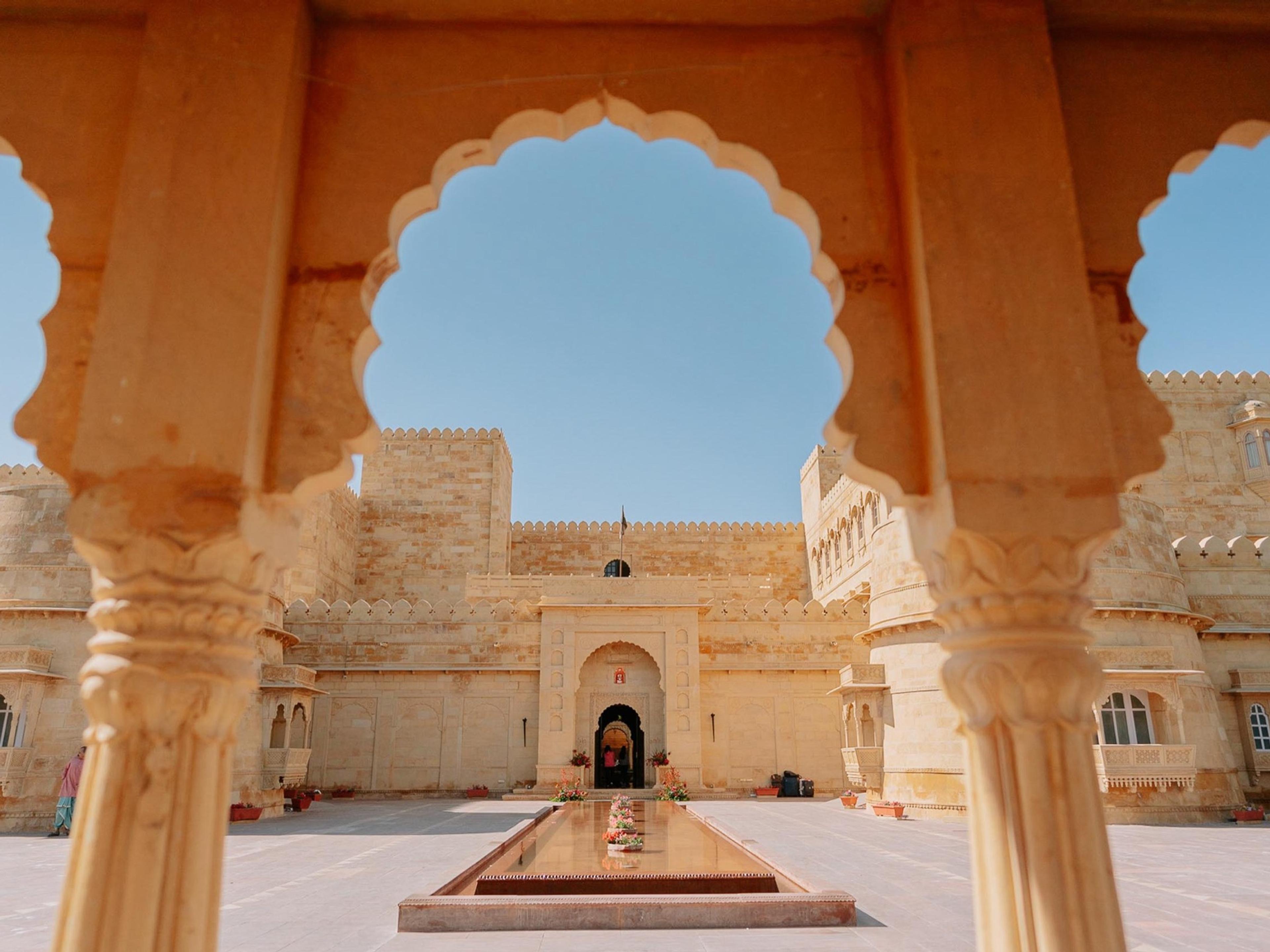 A courtyard of golden sandstone seen through a scalloped arch, with a long water channel to the gate
