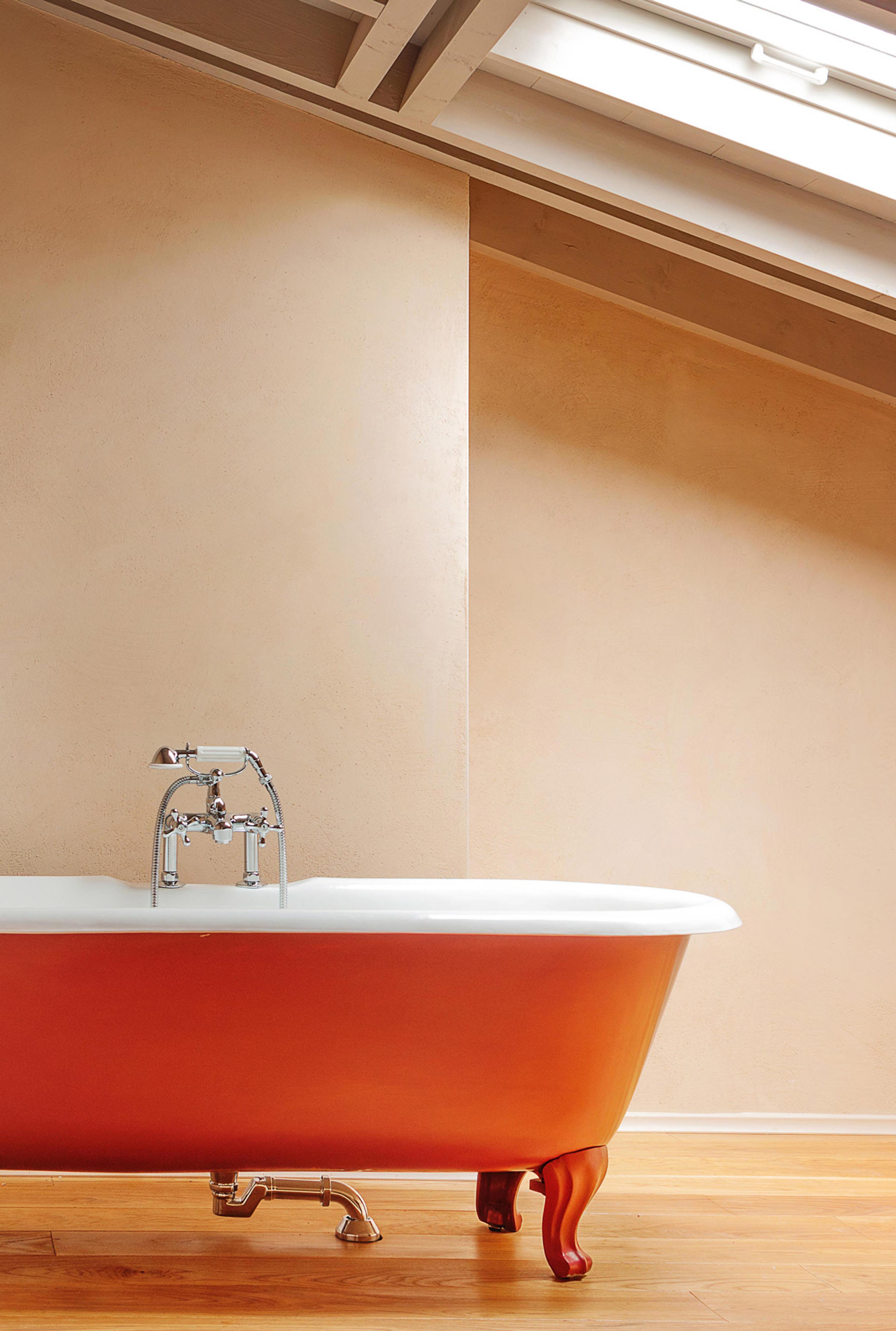 A red clawfoot bathtub with chrome taps standing in a wood-floored attic room lit by a skylight.